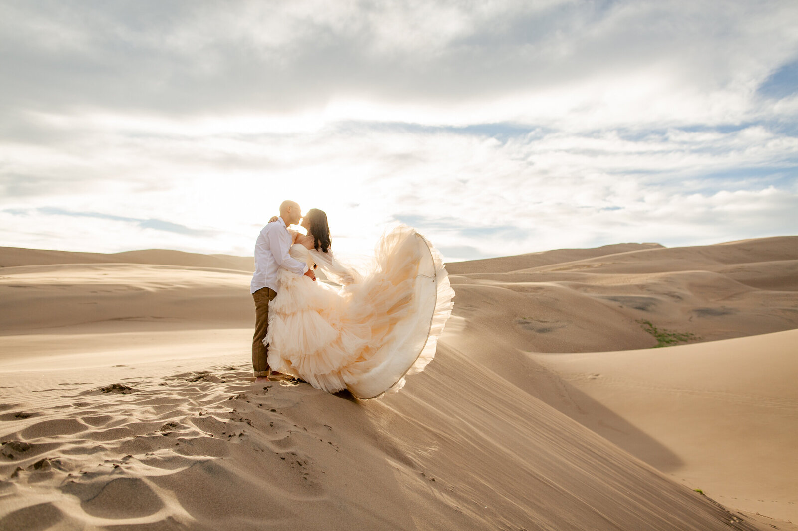 Bride flips up her wedding dress into the wind while hugging the groom on the top of a sand dune