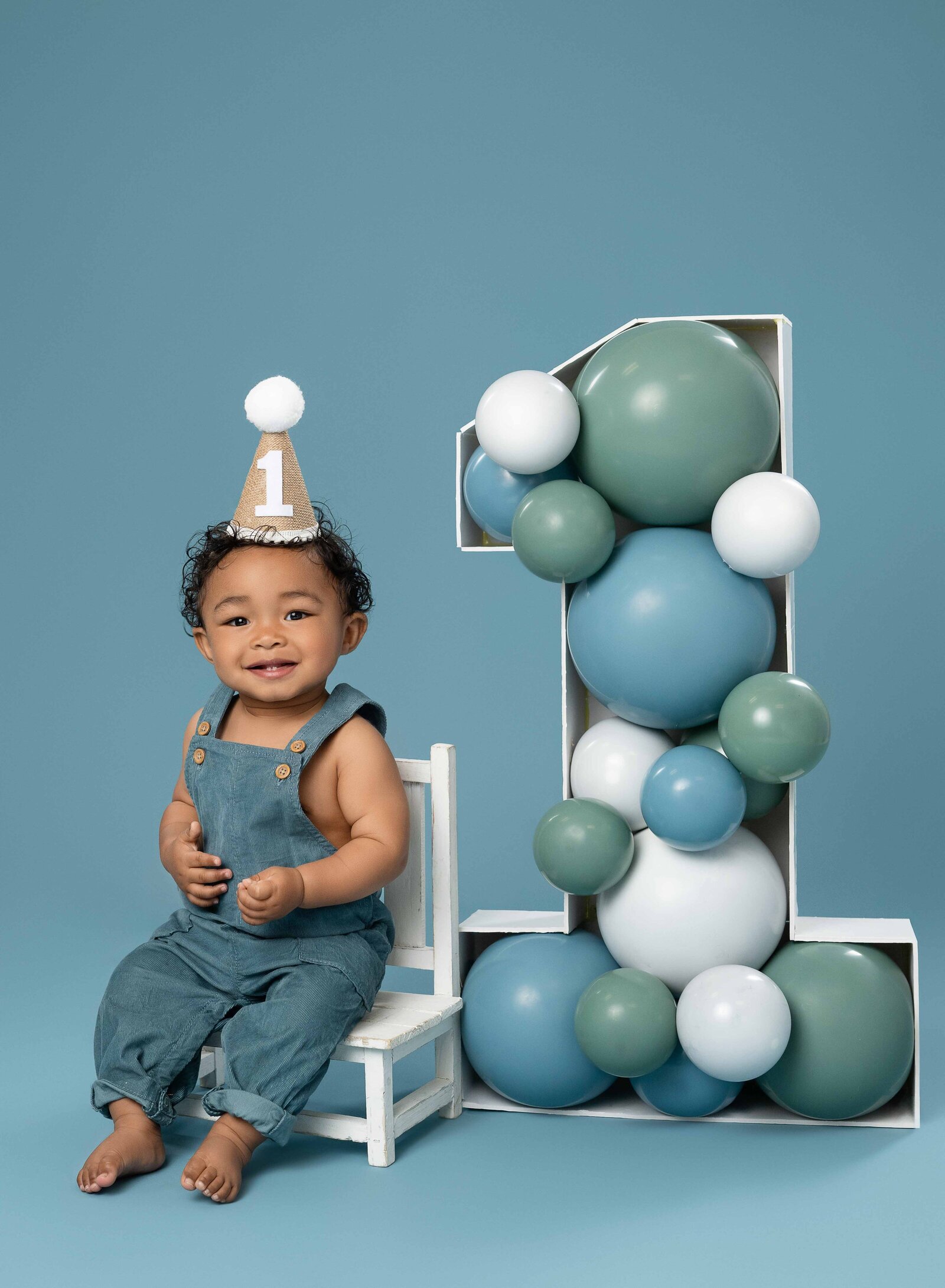 1 year old taking birthday photos, sitting in a chair next to balloon filled marquee, smiling.