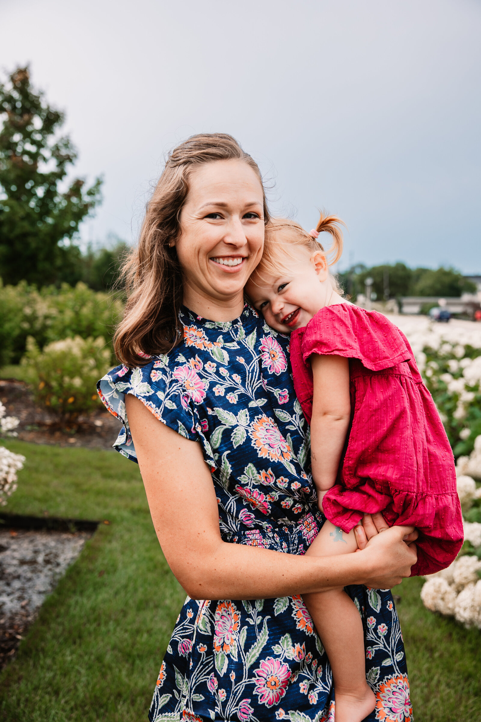 A toddler girl in a pink dress laughs while laying on mom's shoulder in a park garden