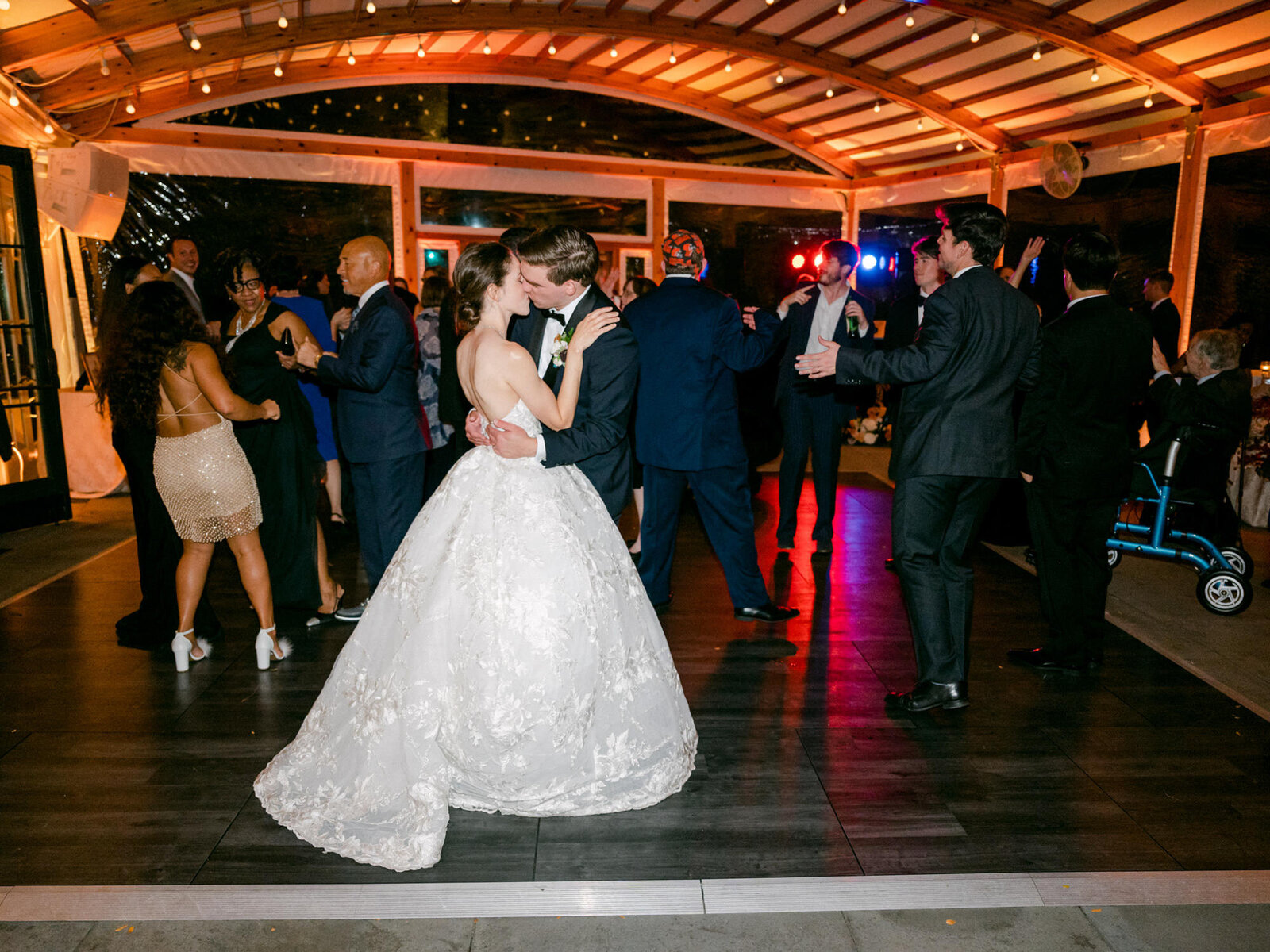 The guests hit the dance floor at a wedding with colorful lights in the background. Bride and Groom kiss in the foreground.