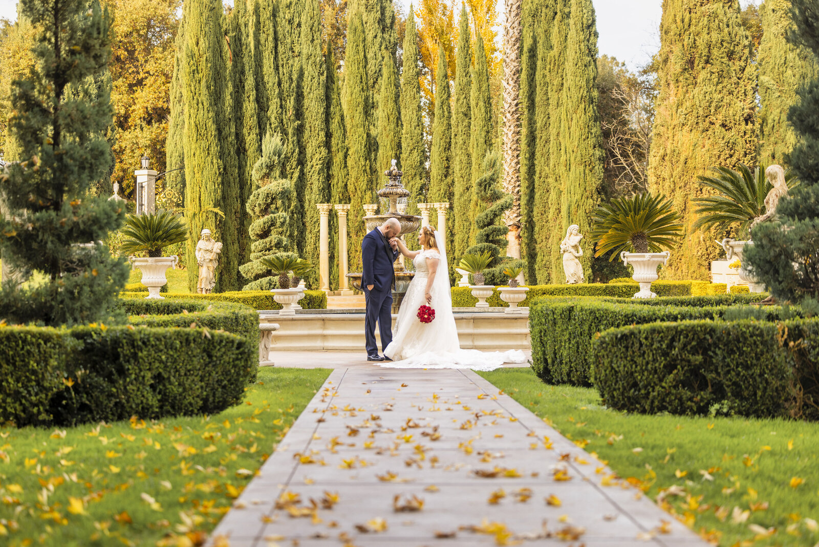 Groom kisses bride's hand at the end of a walkway in front of fountains and large green landscaping at Grand Island Mansion. Photo by Grand Island Mansion preferred photographer in Sacramento, Philippe Studio Pro.
