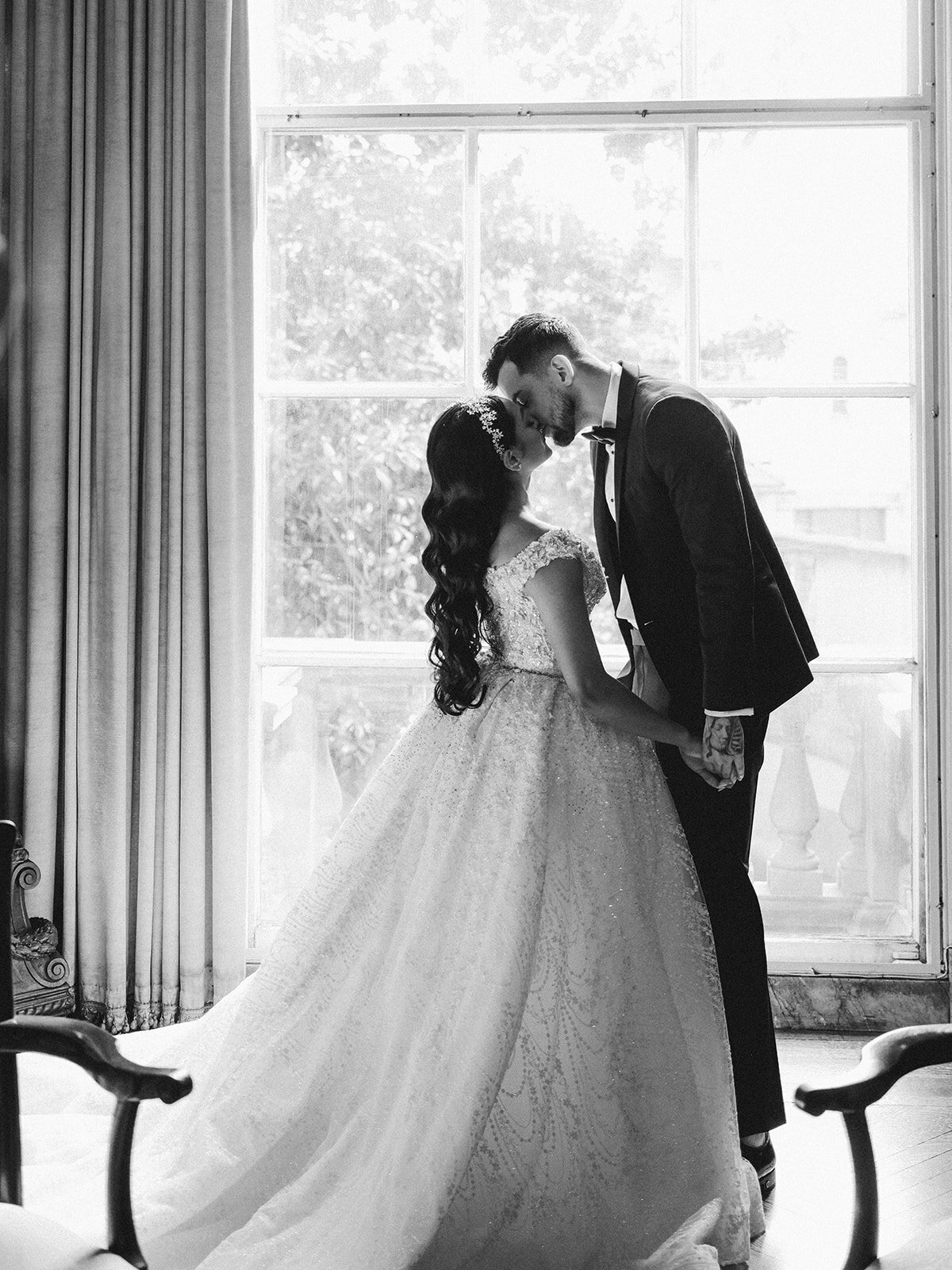 Bride and groom share a kiss in front of a window flooded with light captured in black and white