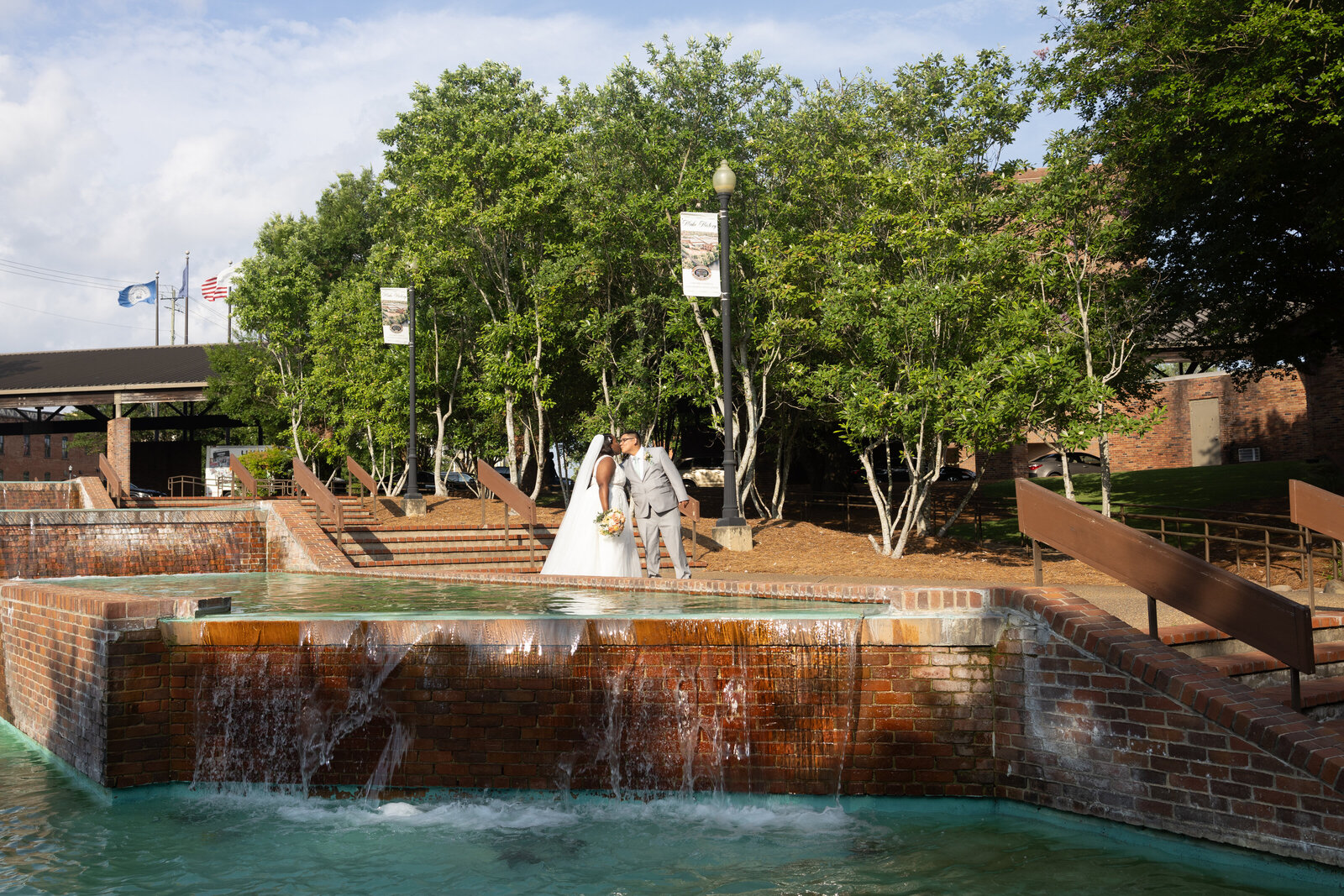 bride and groom kissing while walking down stairs in front of water fountain with sun shining on them