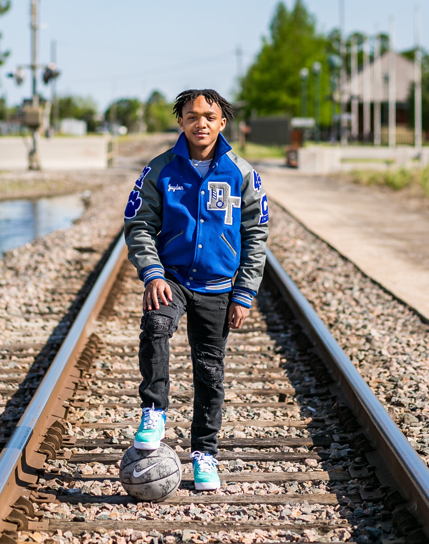 boy with blue letterman's jacket standing on between railroad tracks with one foot on basketball