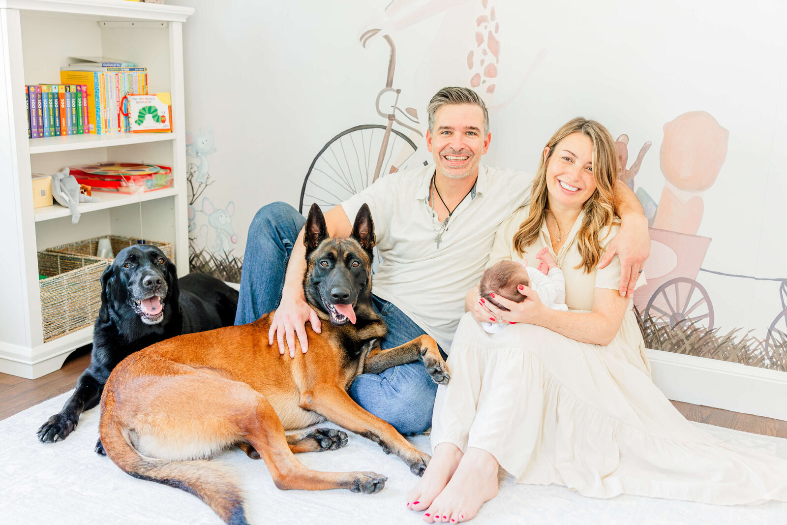 Mom and dad and both dogs smile and/or look toward the camera while mom holds newborn