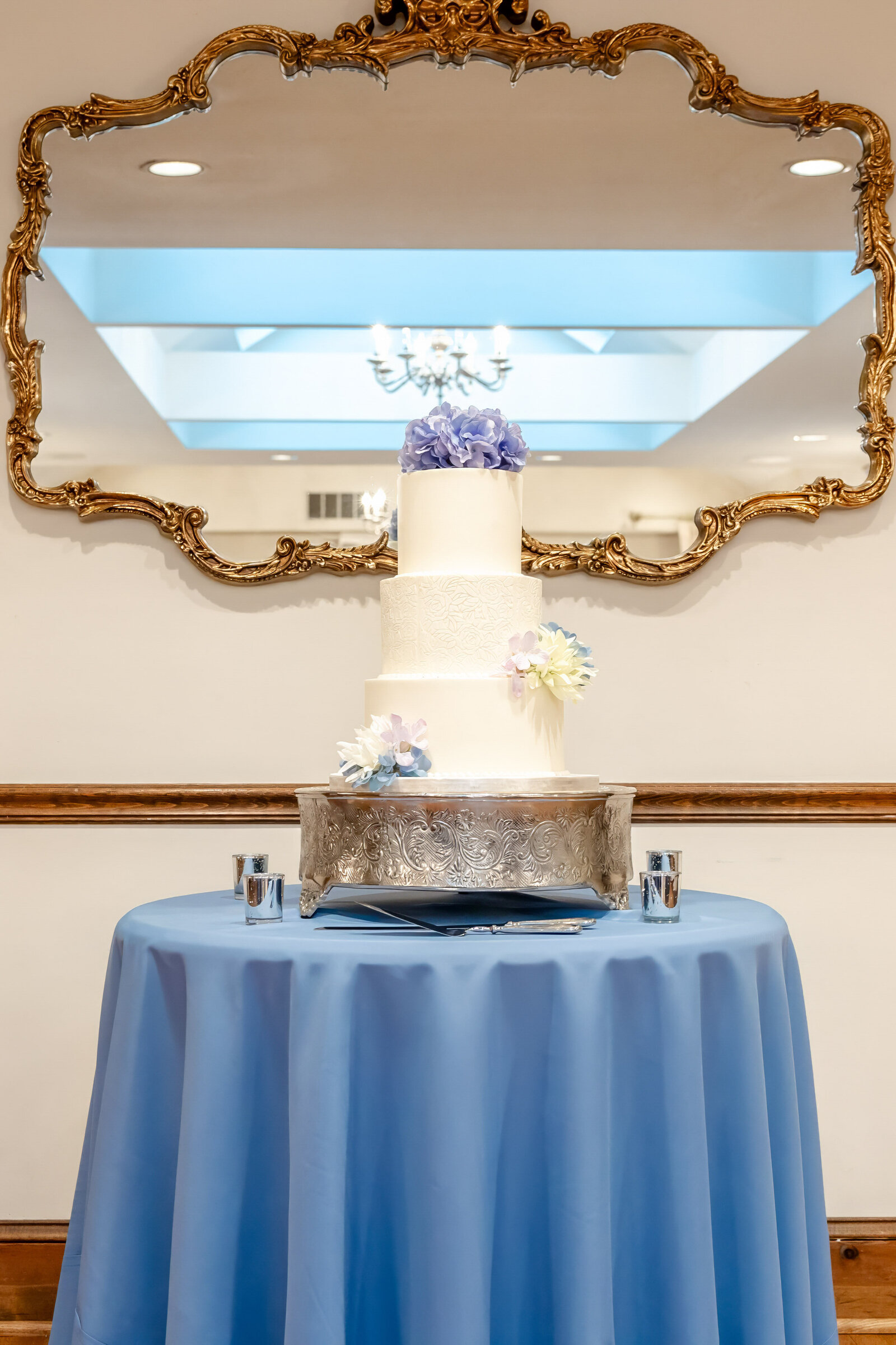 A three-tiered wedding cake decorated with soft purple and blue florals sits on a silver stand, displayed on a blue tablecloth with a gold mirror in the background, captured during a Wilmington, NC wedding.
