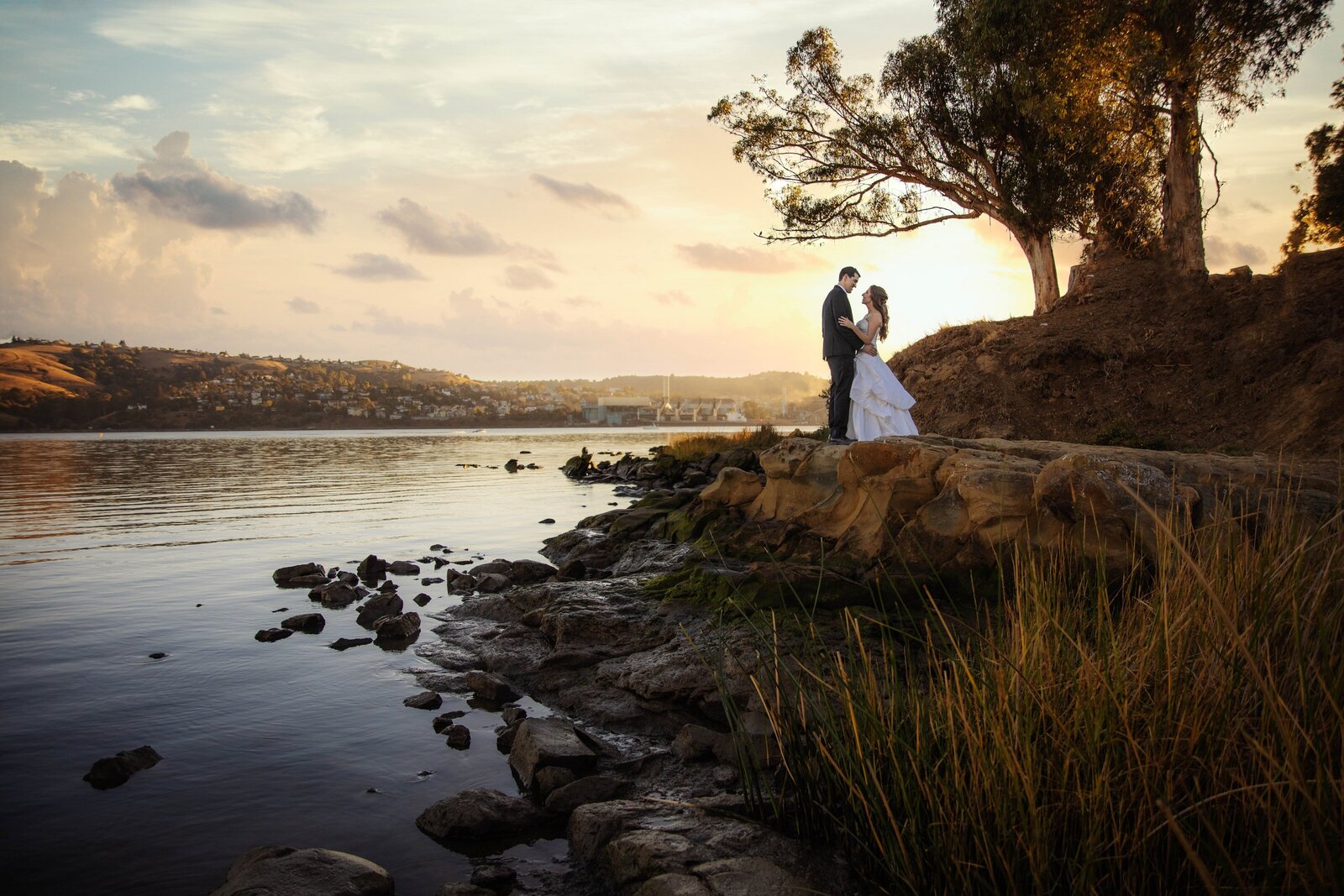 Bride and groom look at each other standing on a ledge that leads into rocks and a river as the sun sets. Photo by wedding photography studio, philippe studio pro.