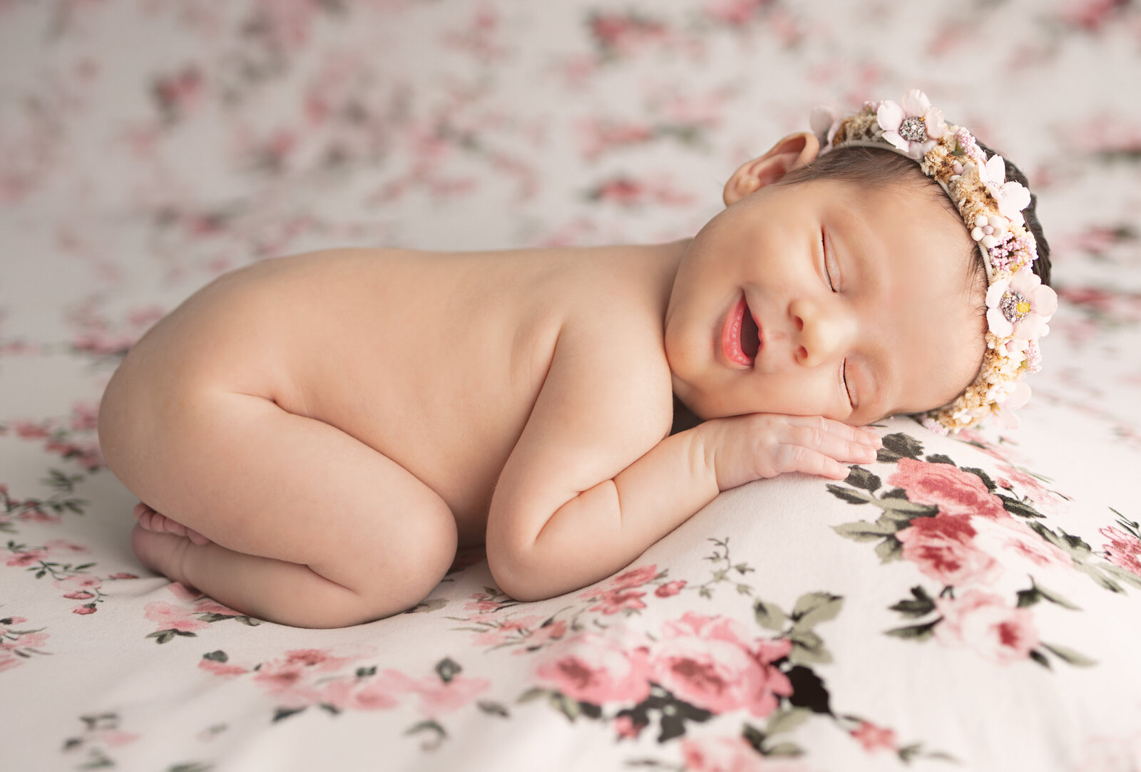 Newborn baby girl smiling in floral headband posed on pink floral blanket during professional Waldwick NJ photoshoot