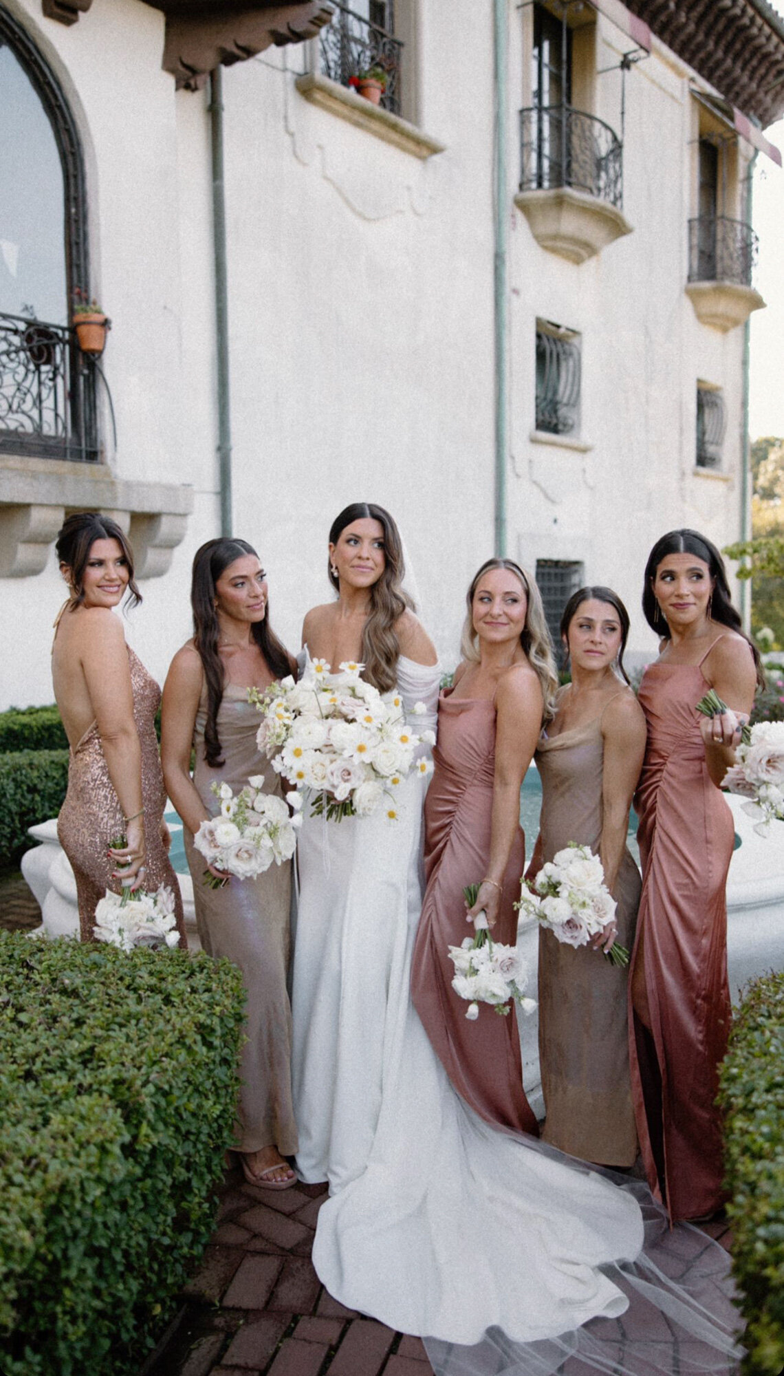 bride in a white dress poses with five bridesmaids in various shades of shimmering dresses, holding white flower bouquets
