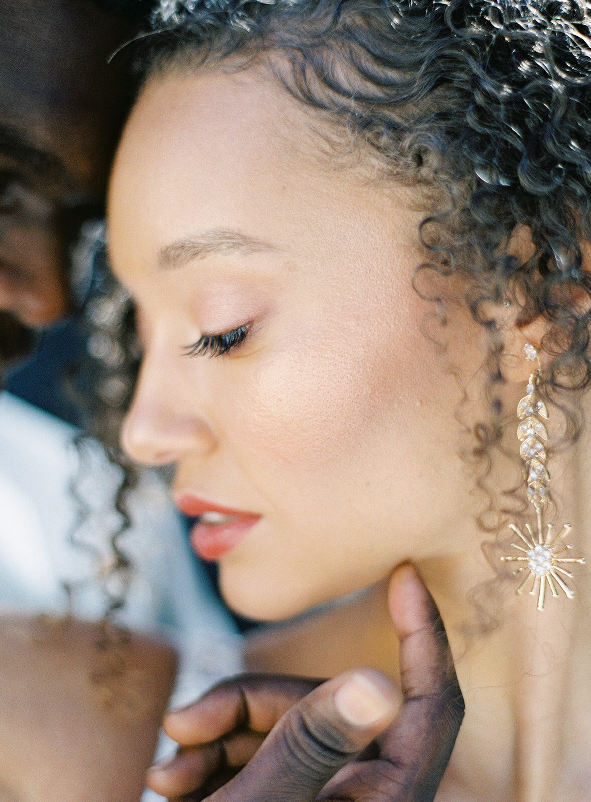 Close up of bride's face. She is looking to the side so her full profile is captured.  She has gold sunburst drop earrings. The groom has his forehead against her temple and is just showing his top lip to top of forehead and his fingers that are softly cupping her under her chin. She has bold scarlet lipstick and her hair is down in curls to the side. The light is shining through behind them. Photographed by wedding photographers in Charleston Amy Mulder Photography.