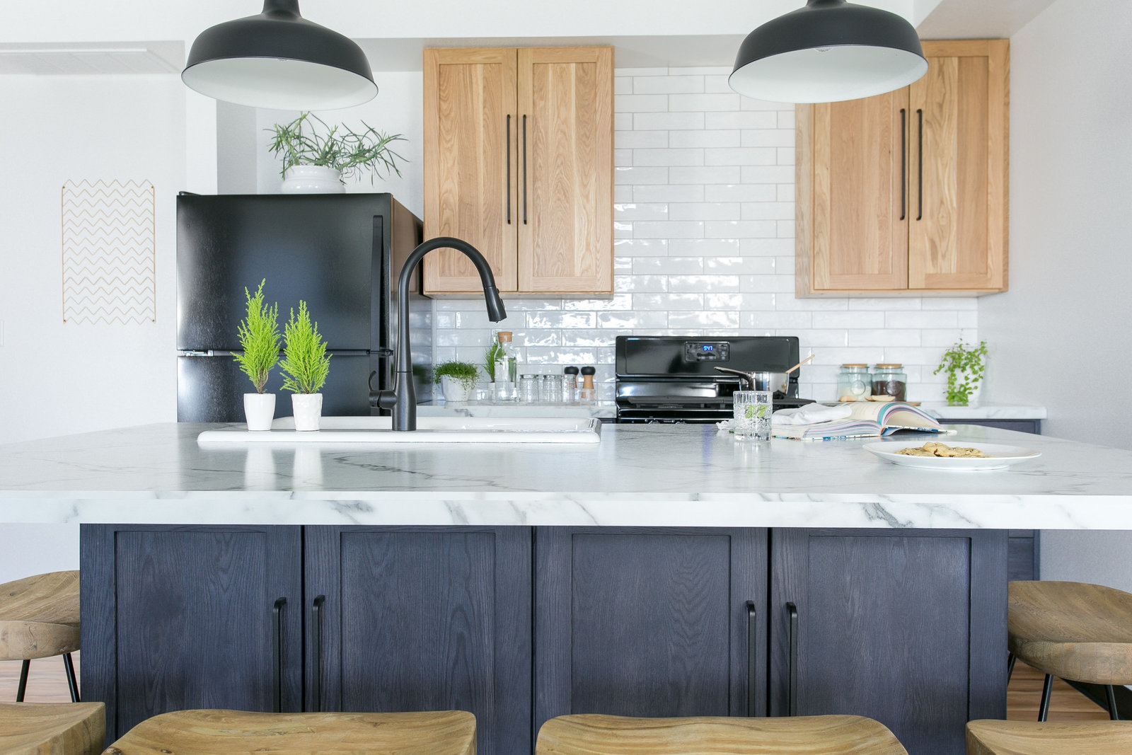 Modern BnB kitchen design with natural oak upper cabinets, navy stained lower cabinets and island base. Minimalist black pendents  with marble like laminate countertops.