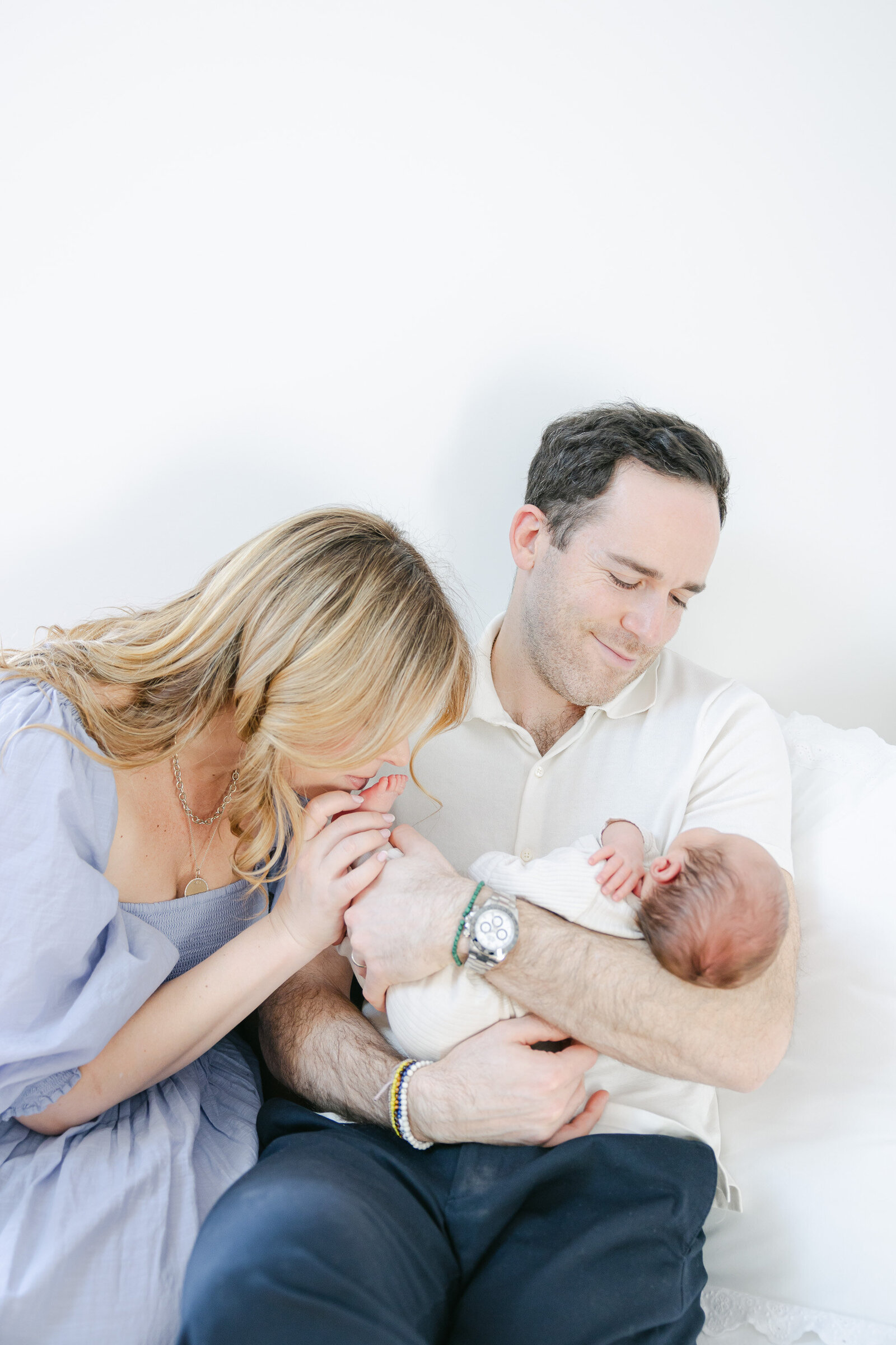 Mother gently kisses her newborn's hand while father cradles the baby with a soft smile during an in-home lifestyle newborn session in Fairfield County, Connecticut. Captured by Nicole Neira Photography, this peaceful family portrait highlights early bonding moments with a natural, light-filled backdrop.