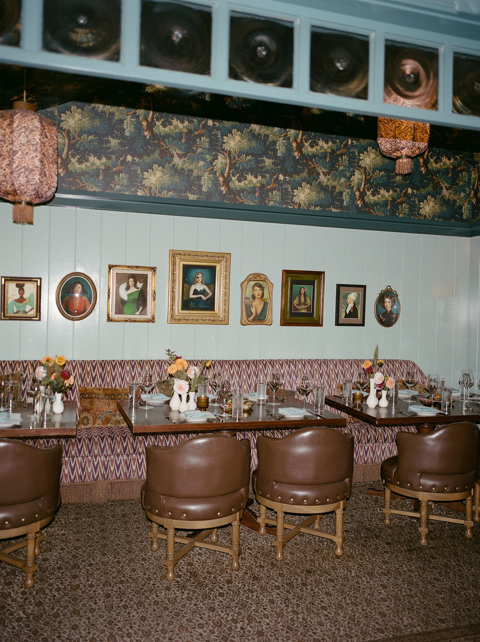 Direct flash film photo of coastal restaurant dining room with teal wall paneling, quirky vintage paintings, and mixed patterns on wallpaper, hanging lanterns, and table booth fabrics 