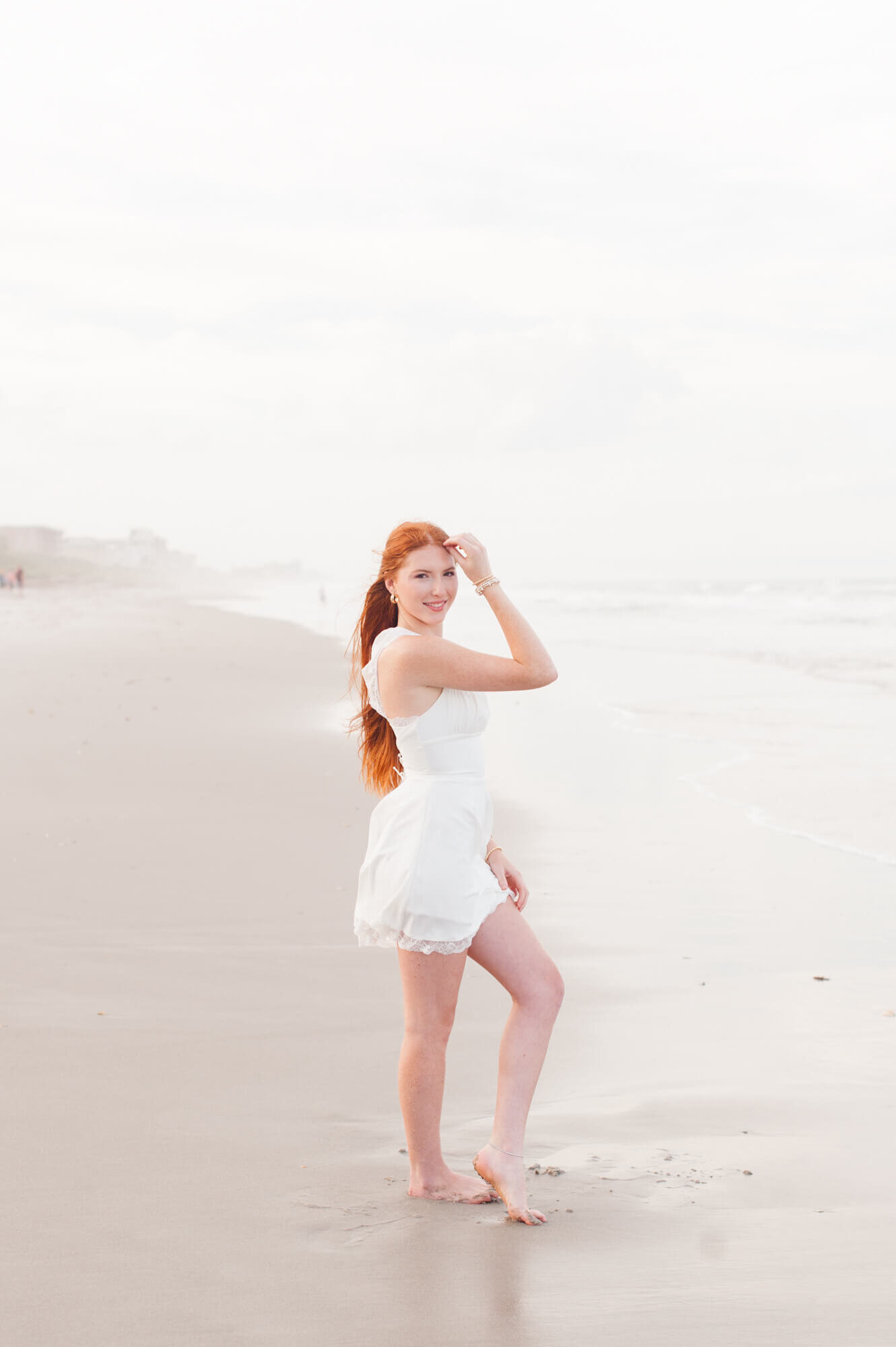 Red headed senior stands on beach in white dress during her senior beach session