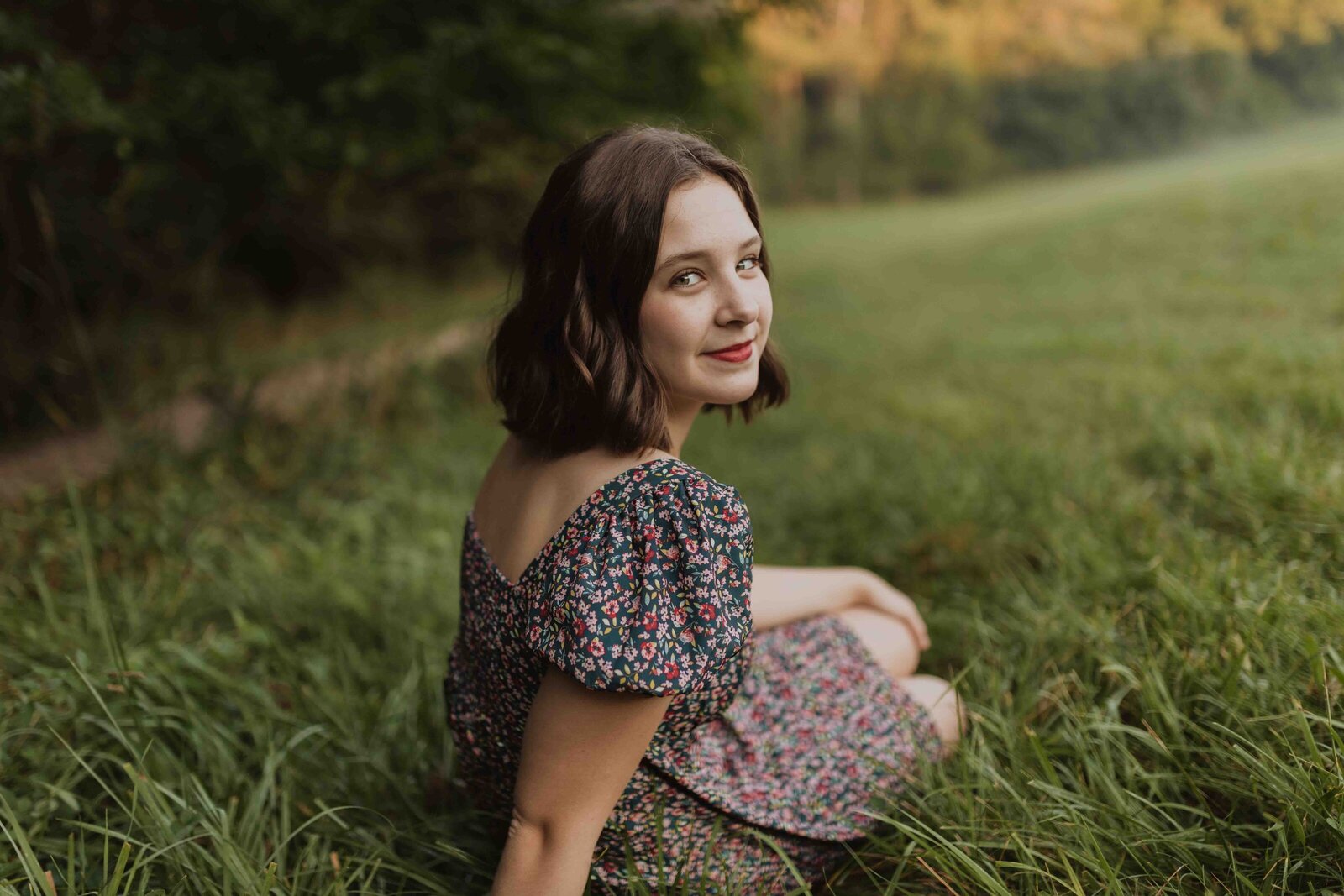 Senior in high school sitting in the middle of field and with her back to the camera and head turn towards camera and smiling at the camera.