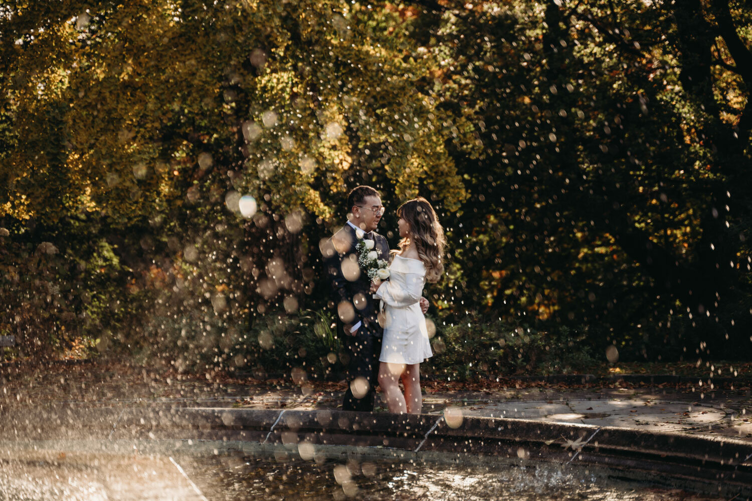 Engaged couple stand behind large fountain as droplets make foreground