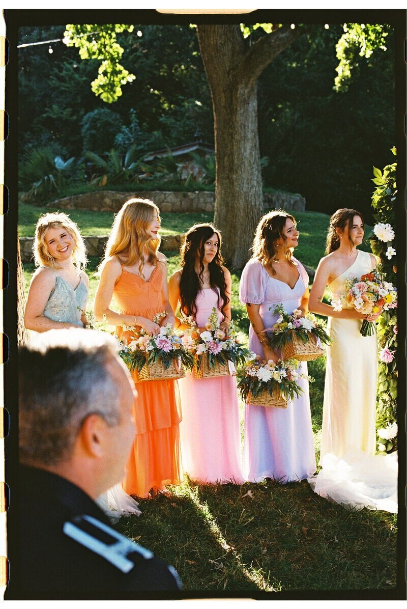 Five bridesmaids in colorful dresses stand outdoors holding bouquets. They are in a sunny, green setting with trees in the background. A man in a dark uniform is partially visible in the foreground.