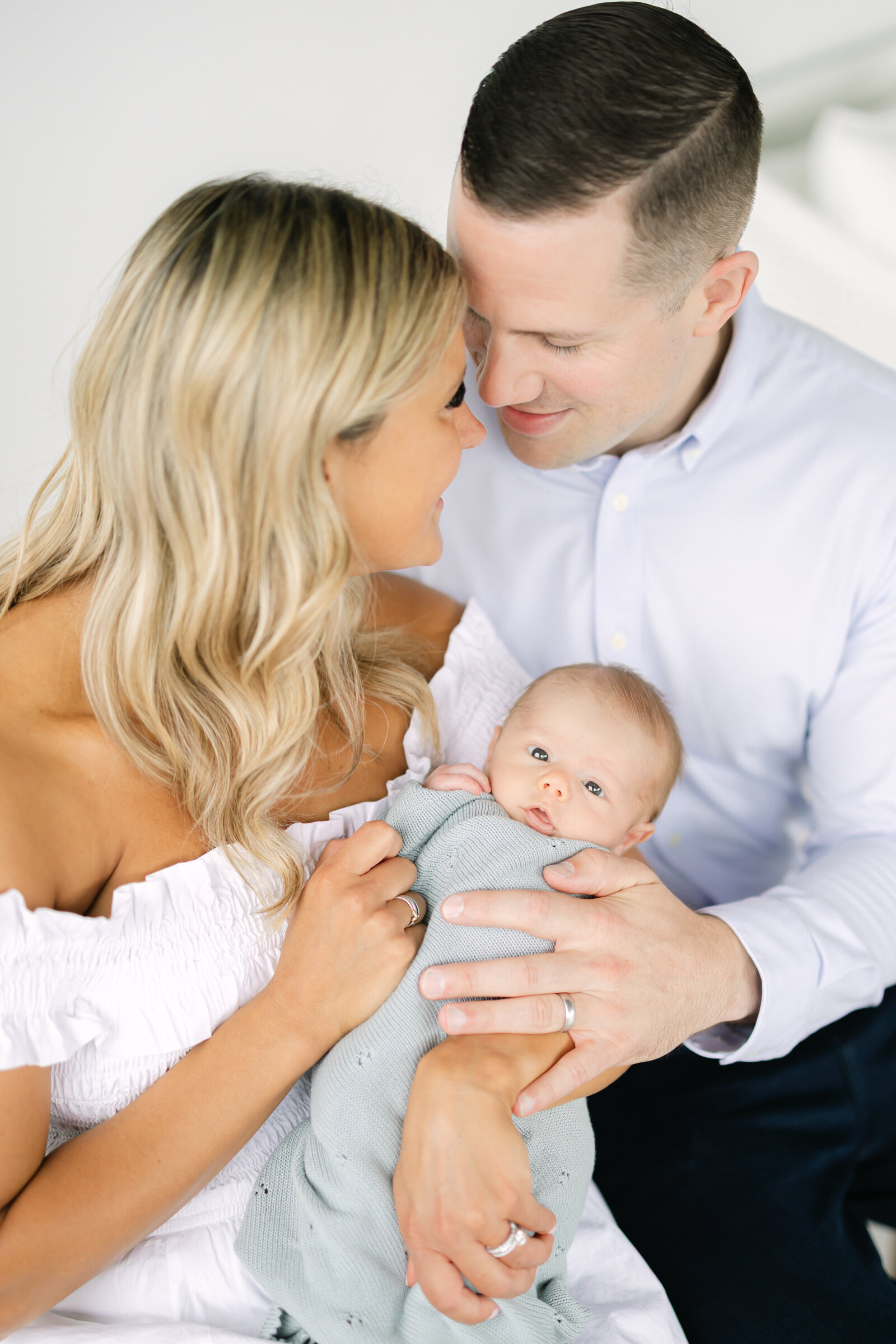 a baby boy wrapped in a light blue blanket is held between his nestling mother and father as he smirks 