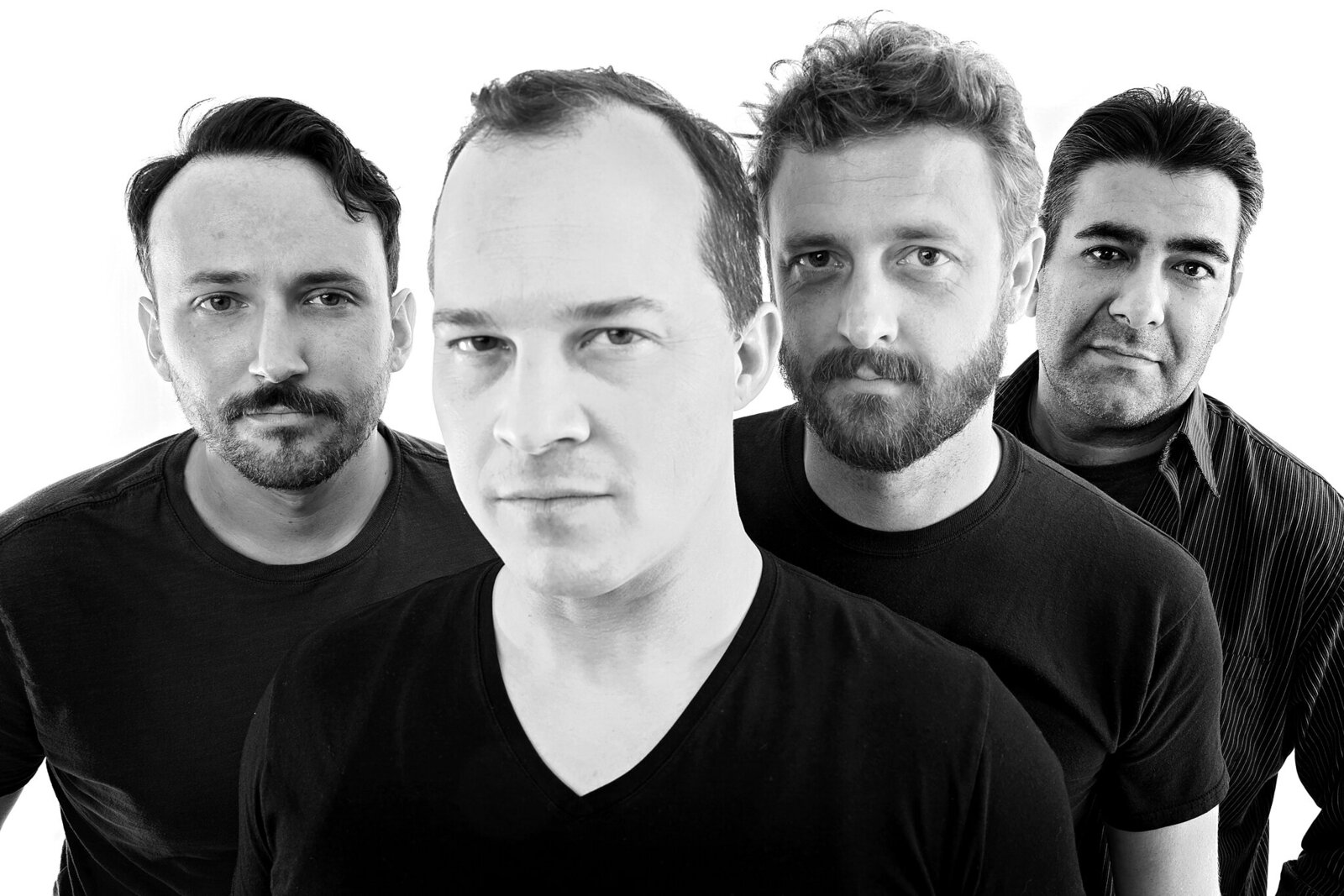 Four men stand in a staggered line facing the camera, all wearing dark shirts. The black and white photo, perfect for professional band photos Jacksonville, features a plain white background and subtle smiles.