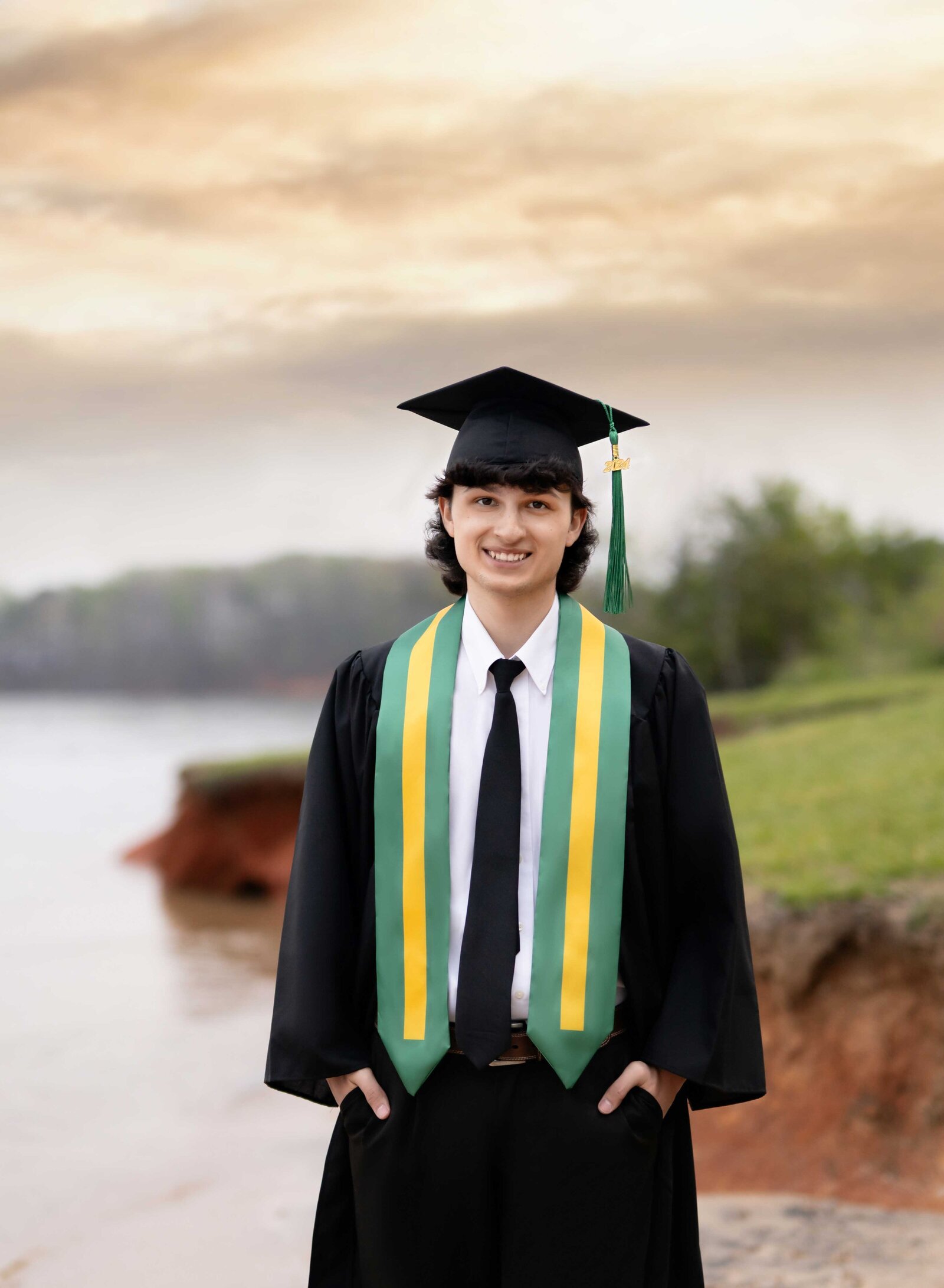 Graduation photo of boy in cap and gown, walking on lake and looking into the camera smiling. Senior graduation photos taken by Jenni Nikki Photography in Atlanta, GA