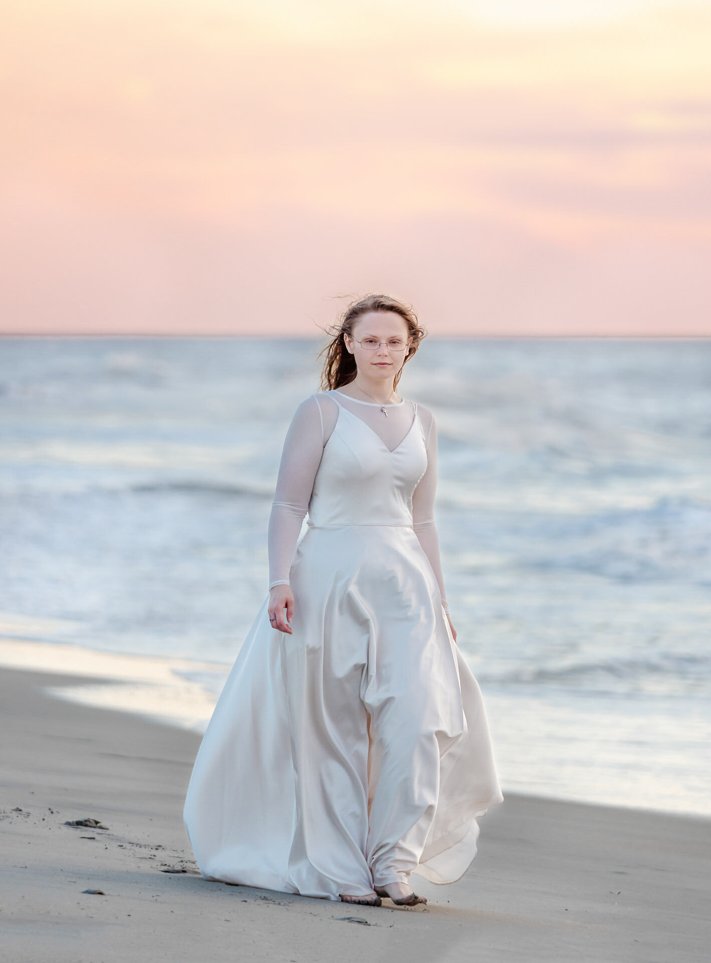 A college senior in a flowing white dress walking along the beach at sunset in Carolina Beach, North Carolina. This serene and elegant image captures the beauty of artistic senior photography, perfect for capturing memorable beach portraits.