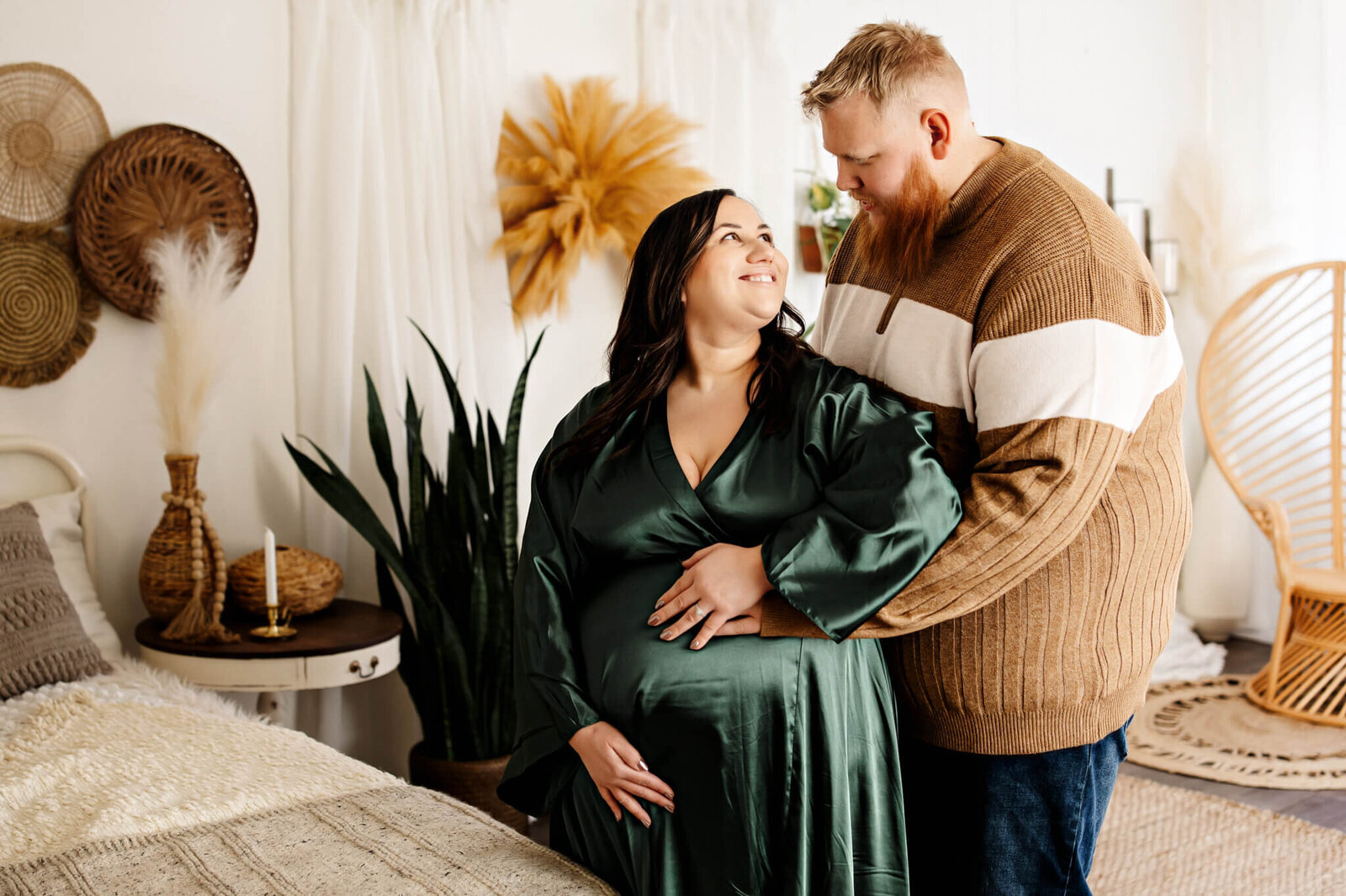 A pregnant woman in a dark green dress stands smiling as her partner hugs her from behind. Perfect for maternity portraits, they share a loving look in a cozy, warmly decorated room with plants and woven wall decor.