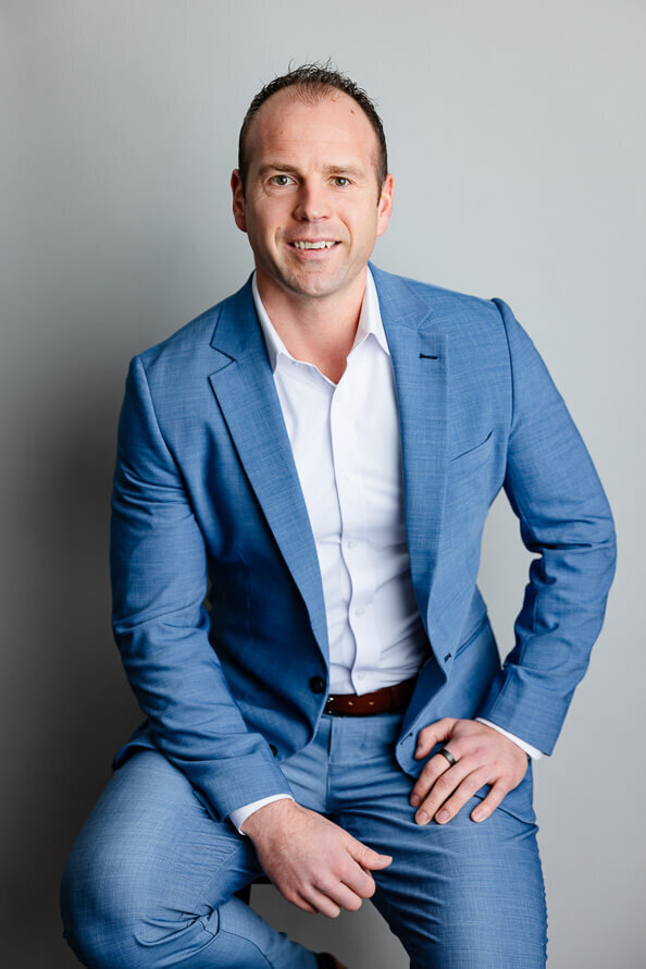 Man in blue suit sits in a gray studio with his hands on his legs.