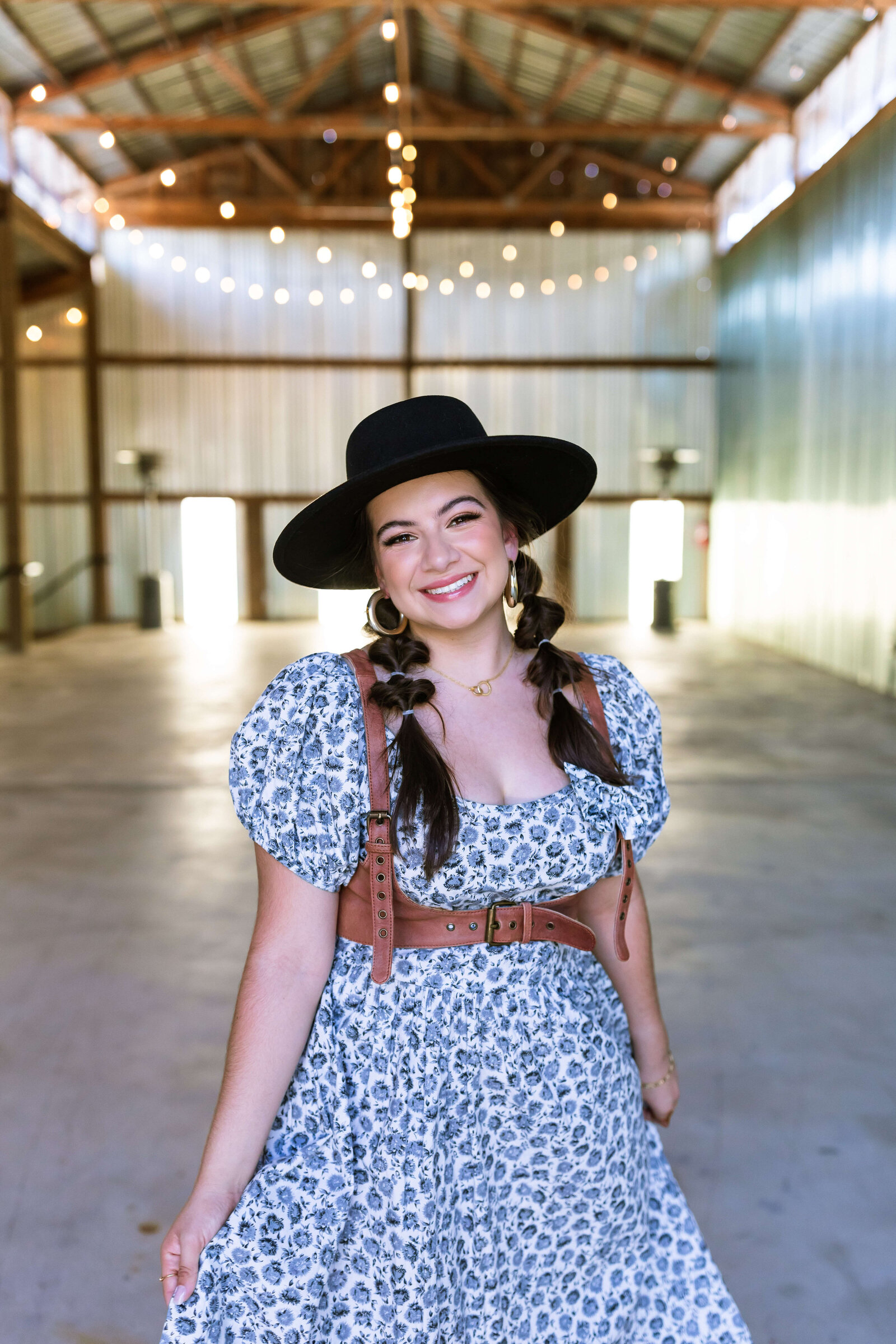 dark haired girl wearing black hat, brown belt, and blue floral dress with bubble braids in a warehouse