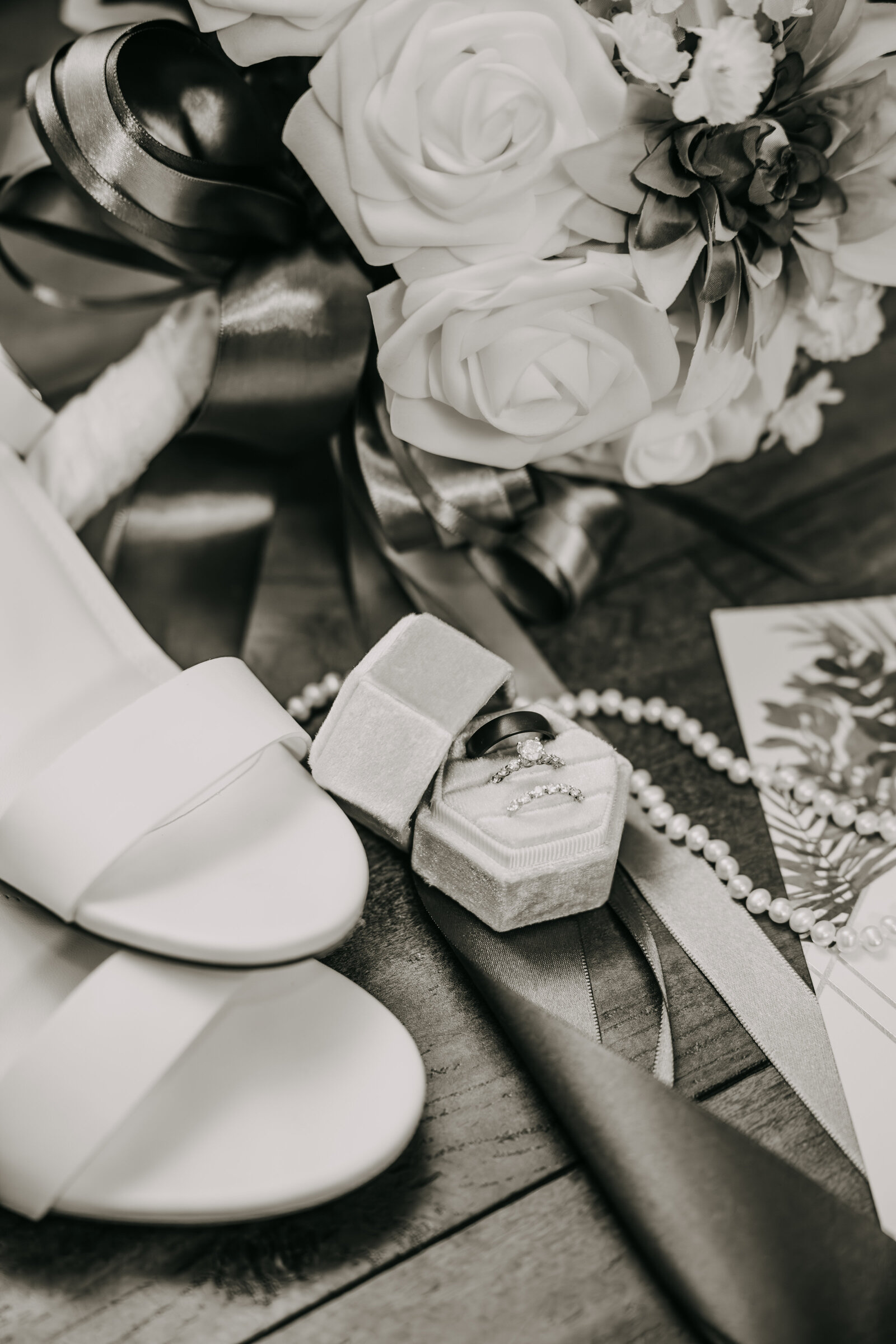 Black and white shot of wedding rings in a velvet ring box, set next to bride's shoes, bouquet, and a string of pearls.