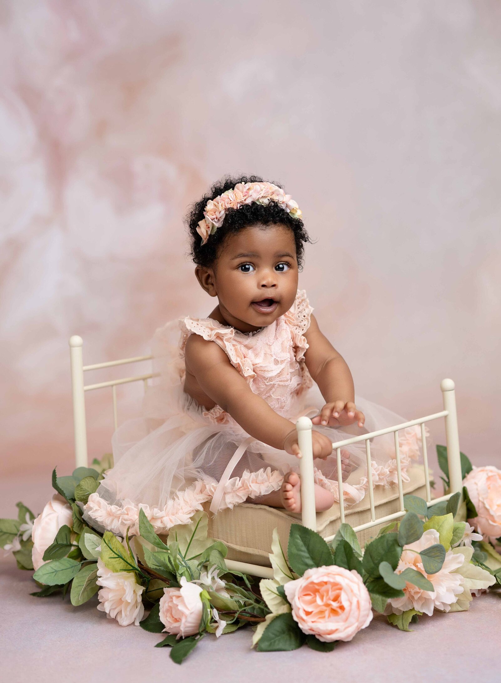 Baby girl sitting on cream bed prop and smiling  with flowers around bed.