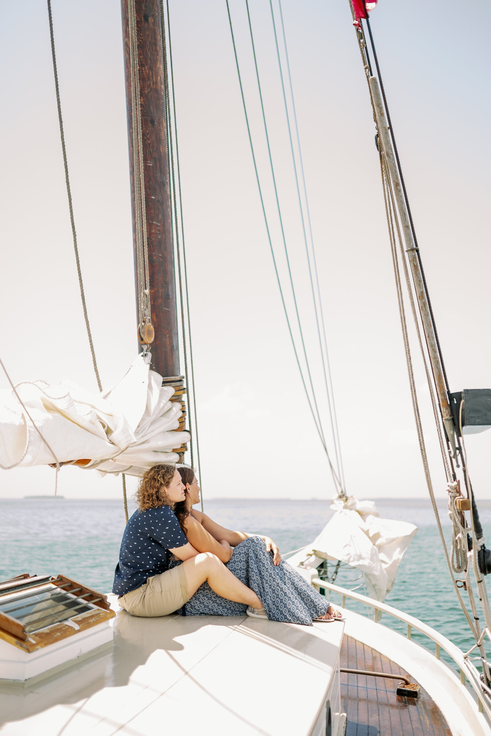 engaged couple sitting on the bow of a sailboat looking out across teal colored water with the sun shining brightly in Key West, FL