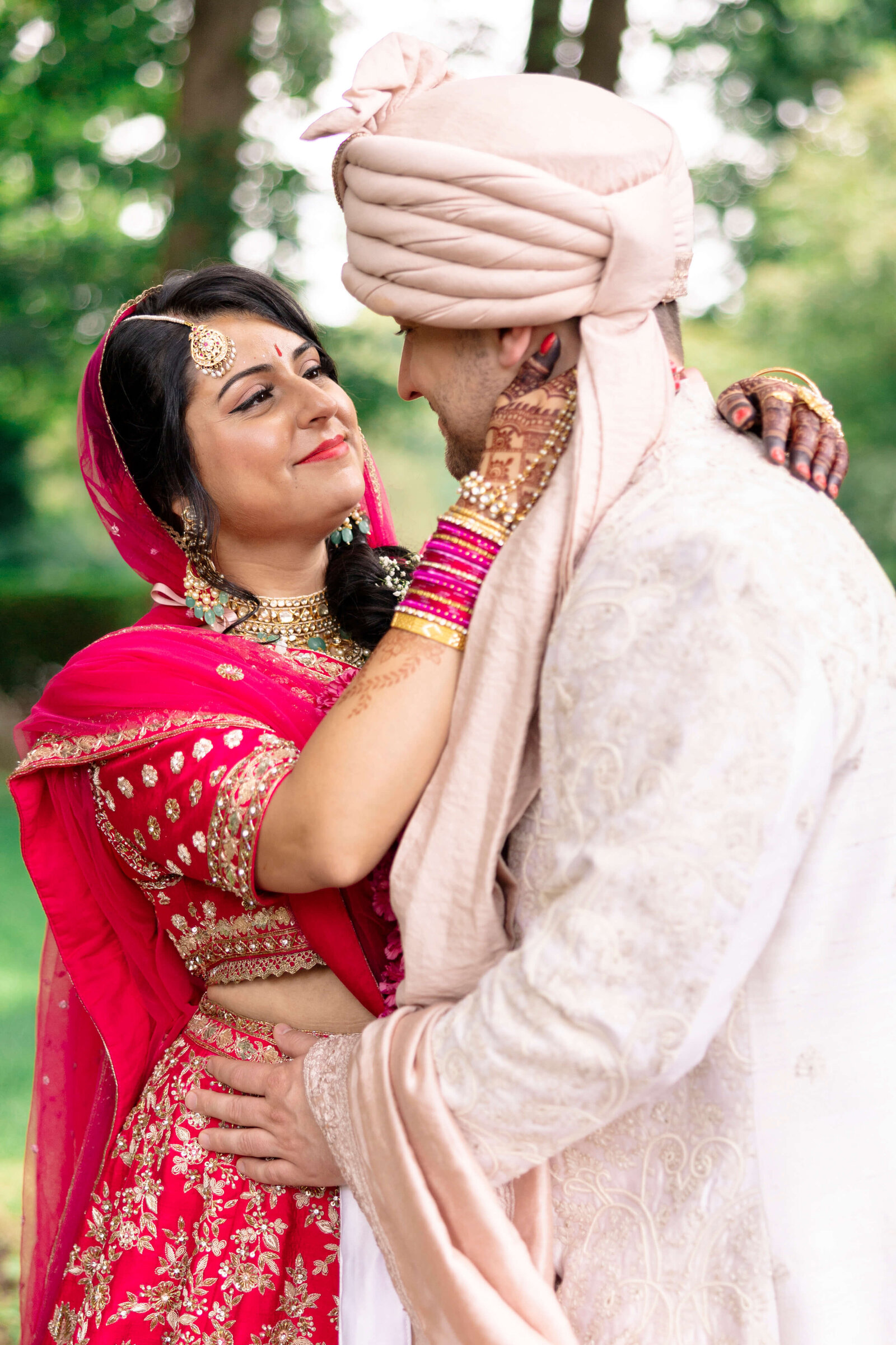 A couple dressed in traditional Indian wedding attire is gazing into each others eyes. The bride is wearing a red lehenga with gold embroidery, and the groom is in a white sherwani with a matching turban. They are outdoors surrounded by greenery.