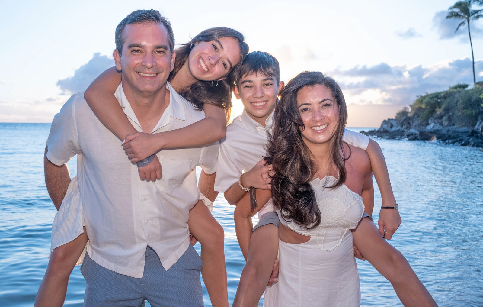 two children piggy back riding their mum and dad on the beach at sunset on their family photo portrait session in Oahu.