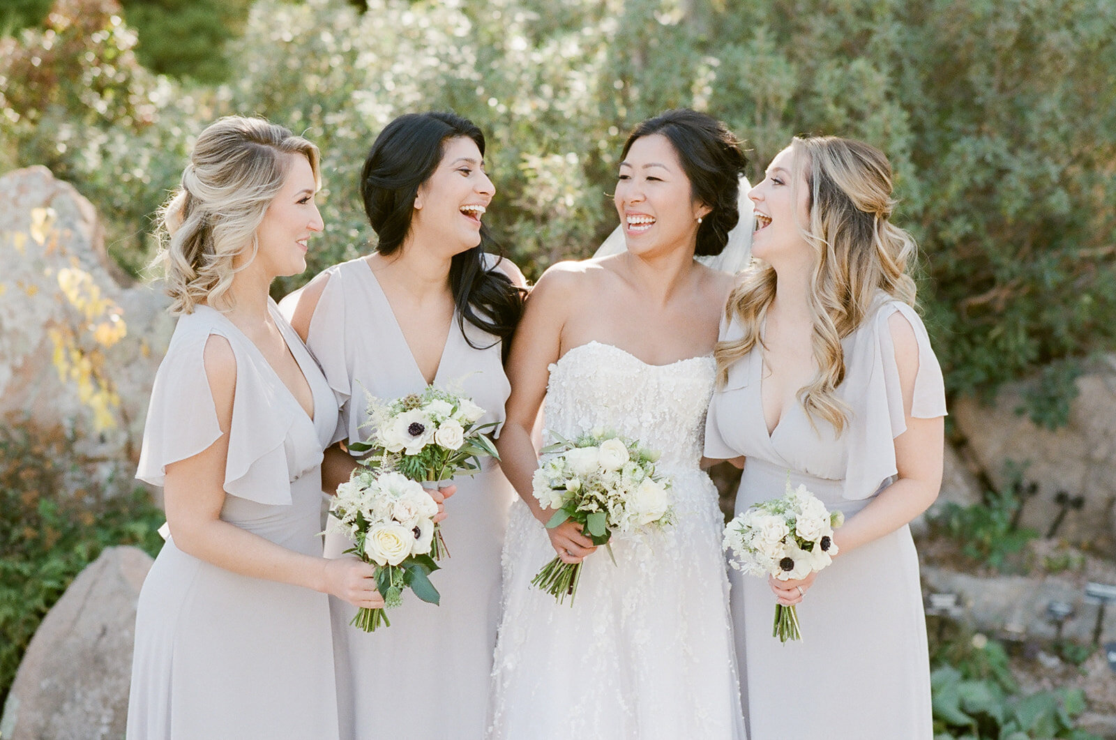 Bride and bridesmaids holding bouquets and laughing in front of trees