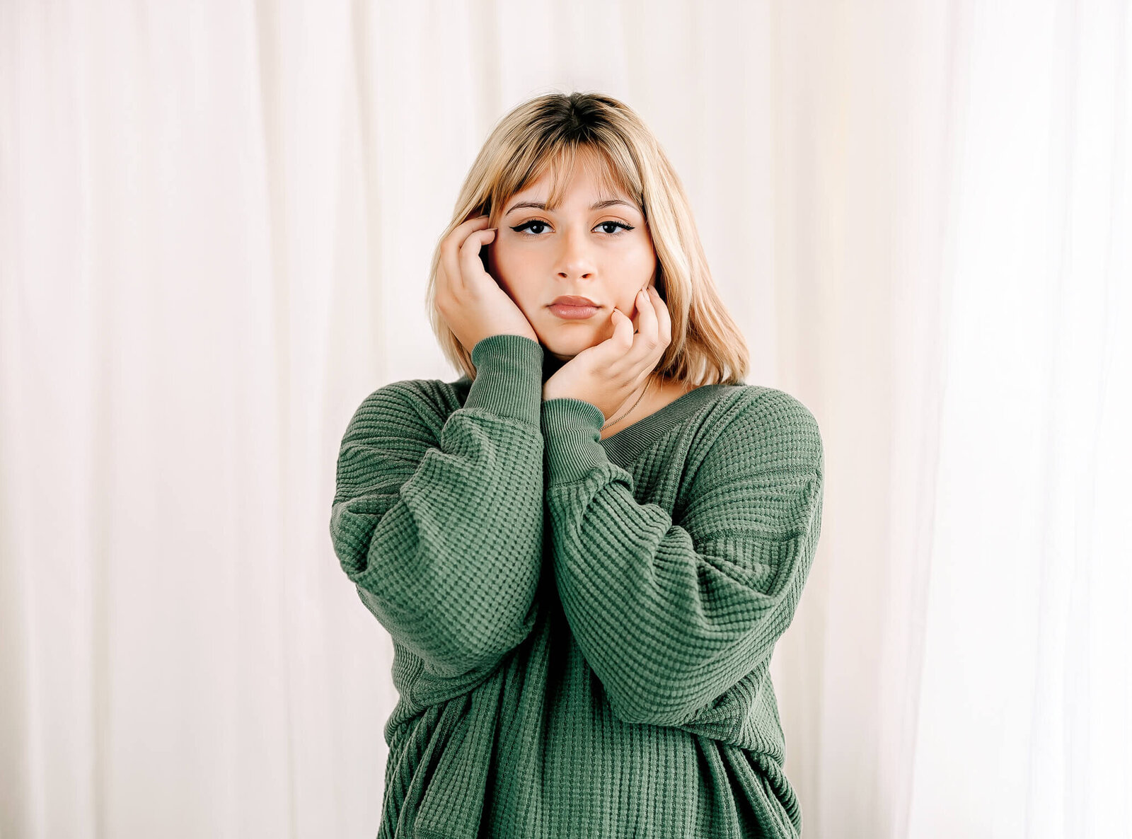 A person with shoulder-length blonde hair wearing a green sweater stands against a white curtain backdrop. They have their hands gently touching their face, looking directly at the camera with a calm expression.