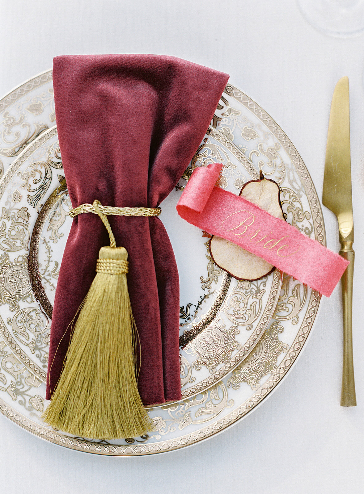 Reception table place setting. Ivory plates with gold intricate detailing. Salad plate is on top of dinner plate with gold brushed knife next to it on the right. Deep red velvet napkin with gold tassel tied around it sit on top of the plate with a dried pear slice next to it with a scroll red tag with gold writing in gold saying 'Bride.' Photographed at Lowndes Grove wedding by wedding photographers in Charleston Amy Mulder Photography.