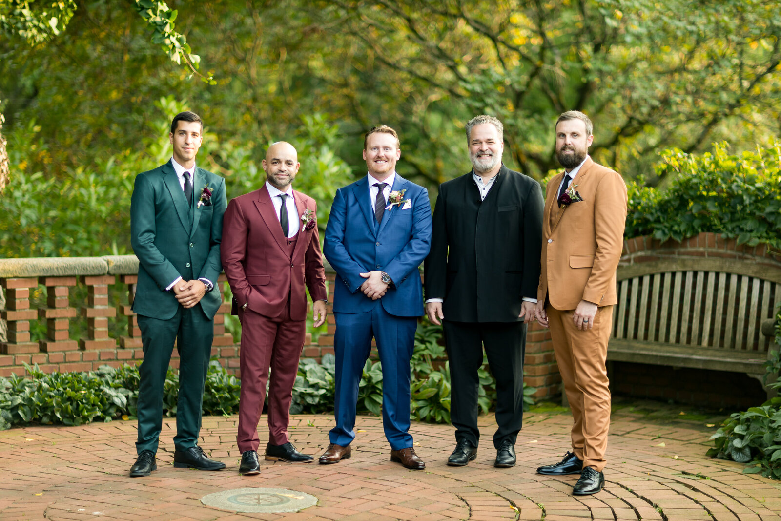 The groom men are standing in a half circle posing around trees during golden hour. The men are wearing fall colors 2024