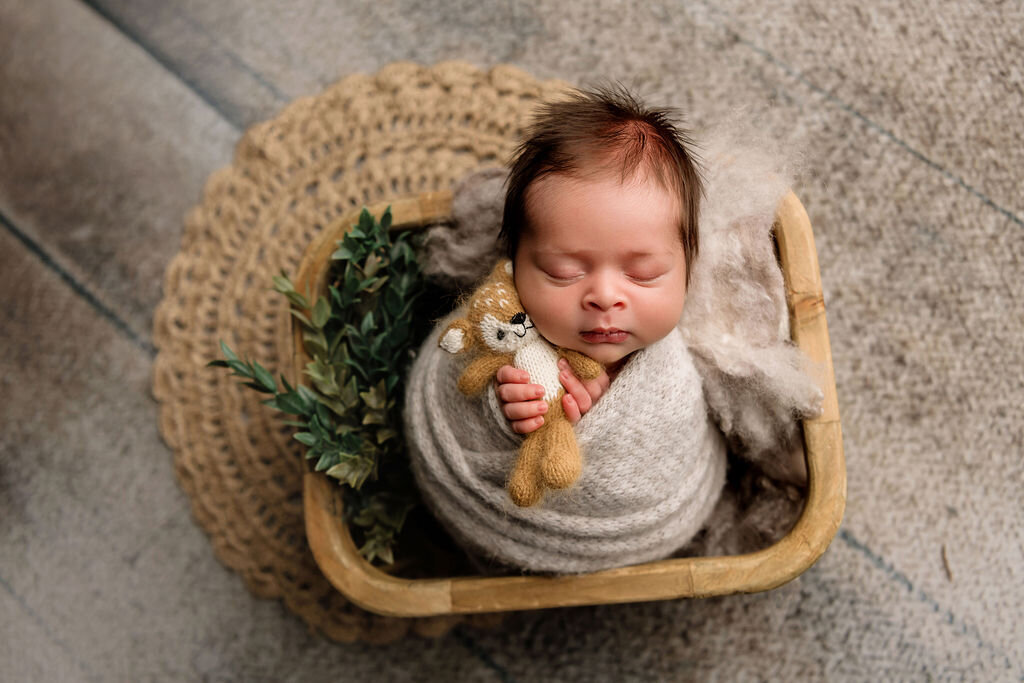 Newborn baby sleeping peacefully in wooden basket while holding small knitted teddy bear