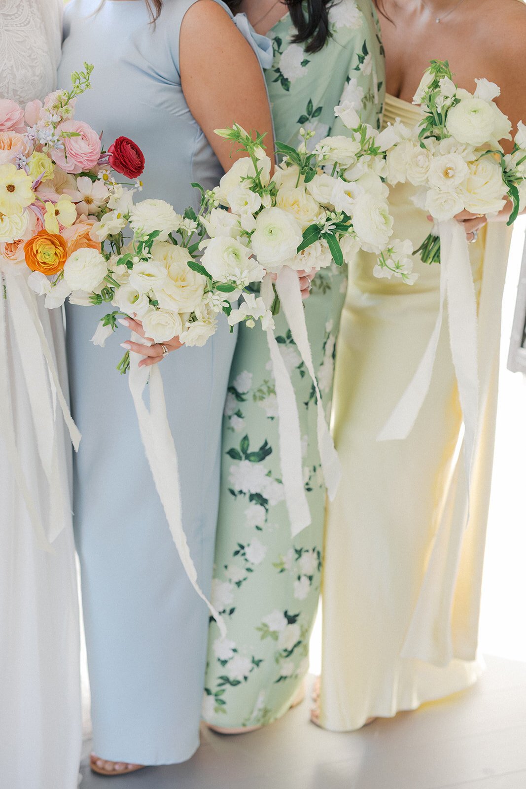 Garden party style bridesmaids is mismatched dresses standing with their bouquets with cream ribbon blowing in breeze from each bouquet. 