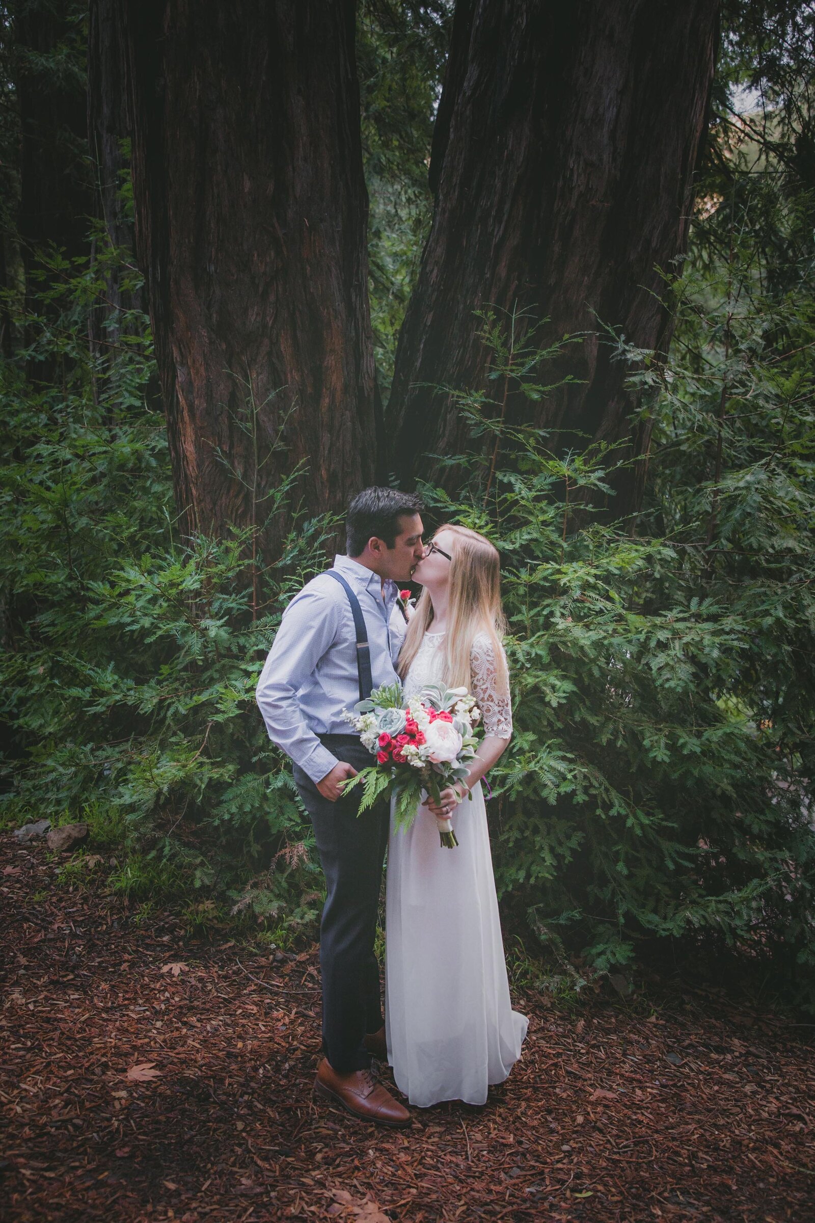 A Couple kisses with redwoods surrounding them.