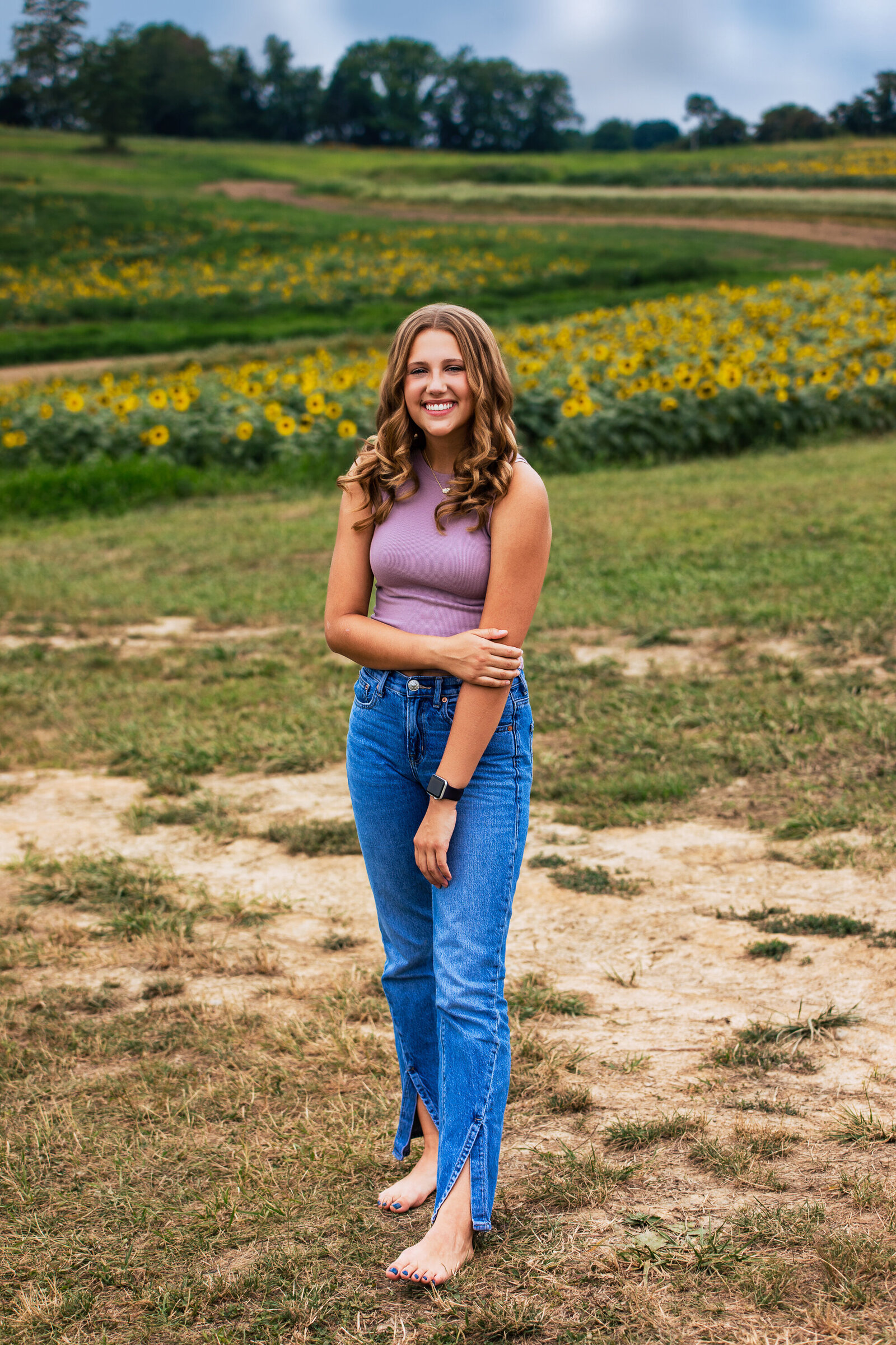 Senior girl poses with a big smile during her senior portrait session in a field with lots of yellow sunflowers near Elizabeth Pennsylvania.