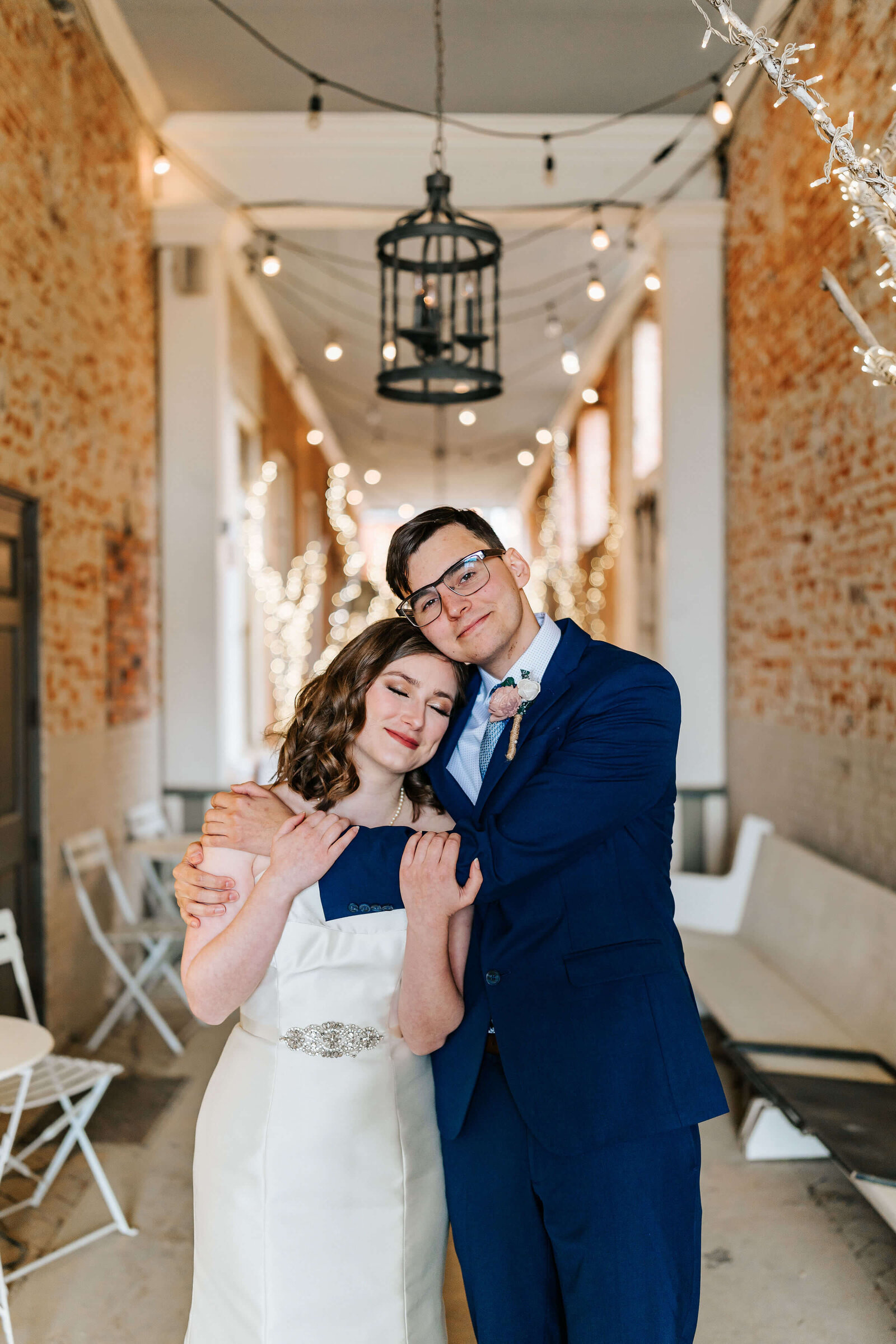 A bride in a white dress and a groom in a blue suit stand close together, smiling and embracing in a decorated, brick-walled venue with string lights overhead.