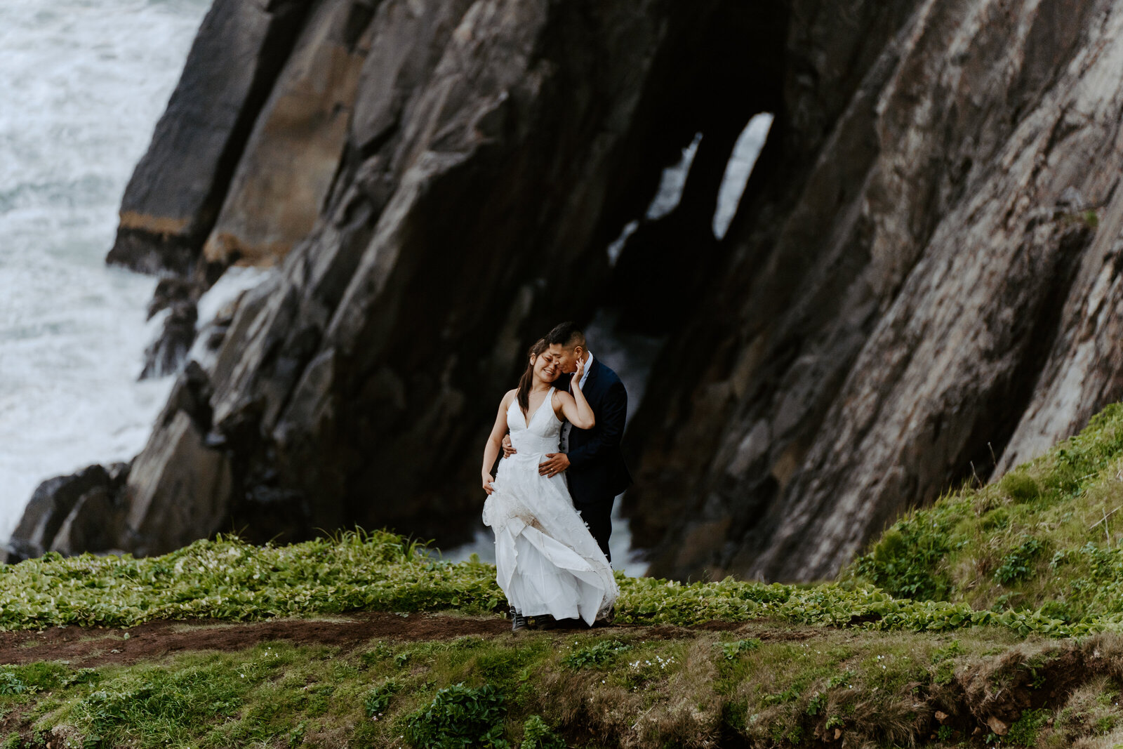 Bride and groom hugging in wedding attire on a grassy hillside with sheer rock cliffs and the Pacific Ocean in the background