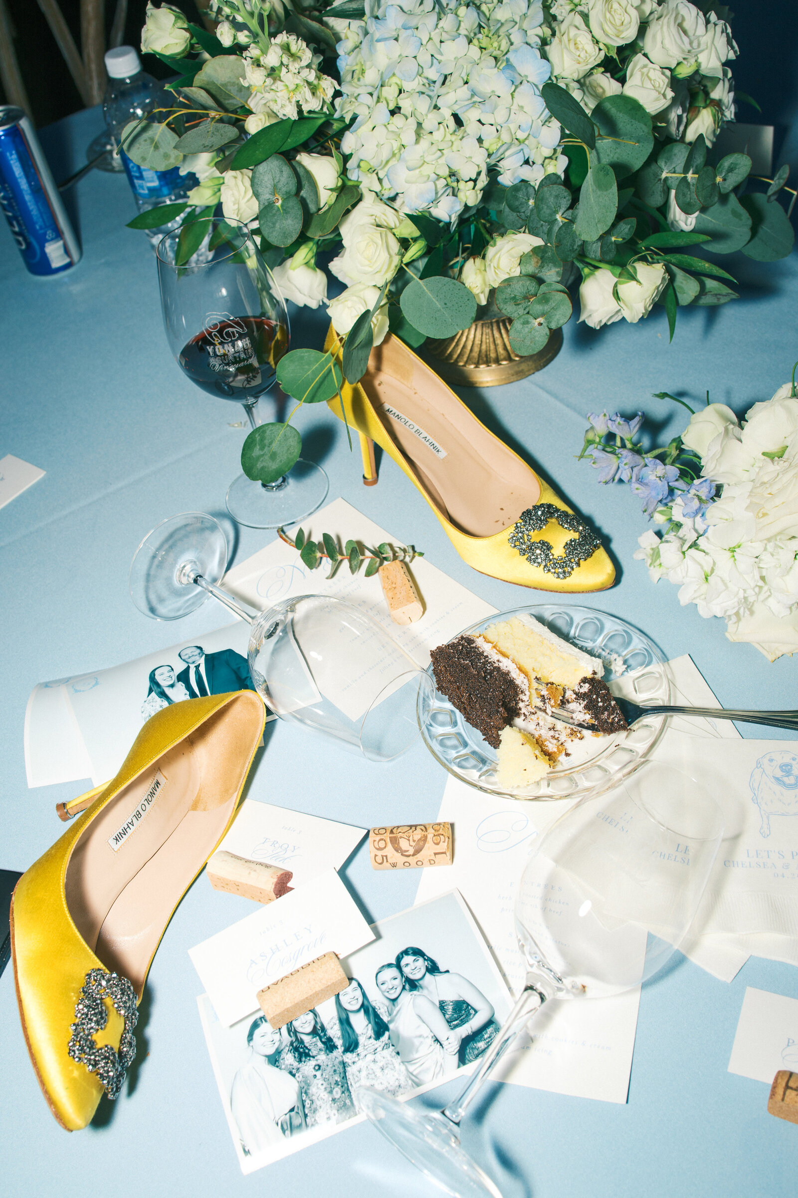 Reception flatlay of Milano heels, piece of cake and wine glasses on blue tablecloth 