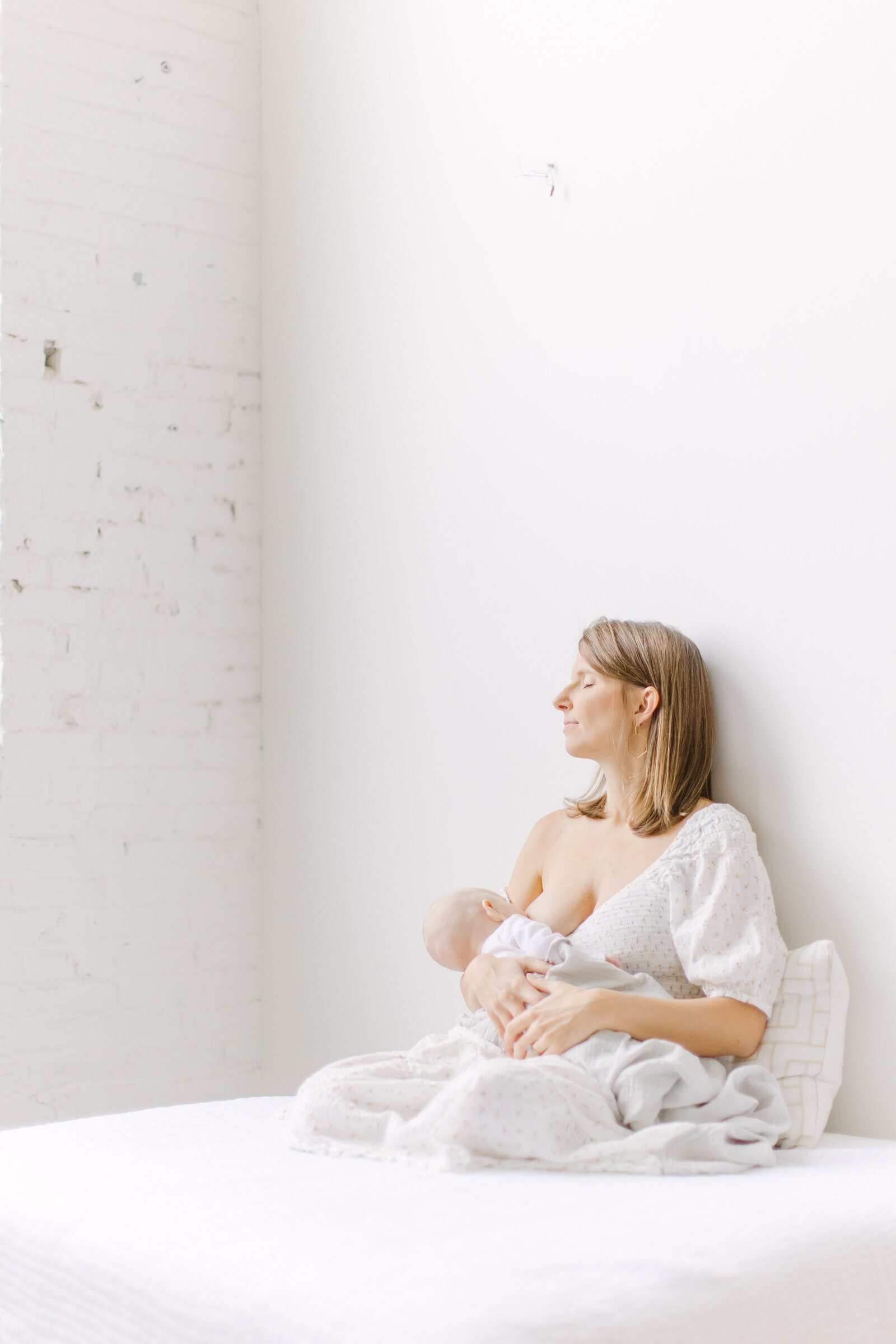 A new mother is breastfeeding her baby while sitting on a white bed in a photo studio in the boston area