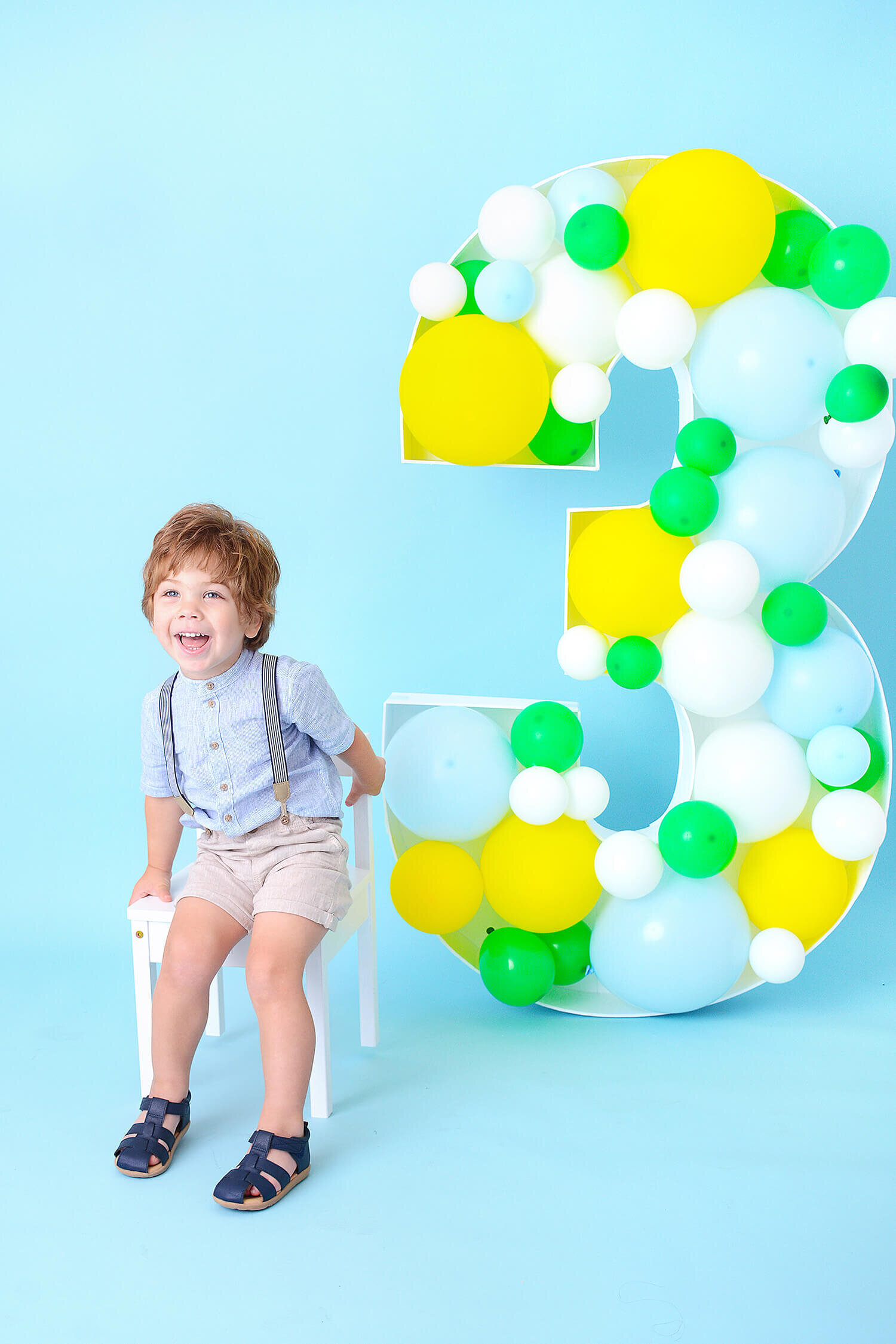 boy smiles at his 3rd birthday photoshoot with a large 3 made from balloons