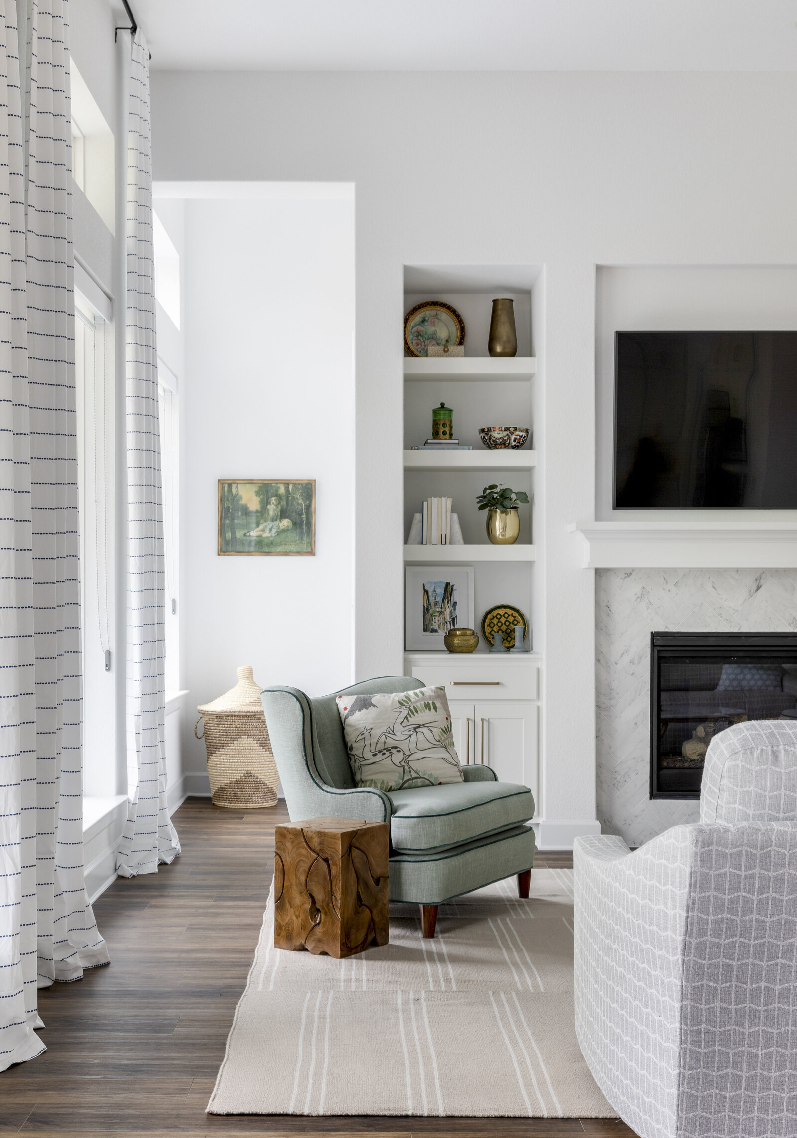white livingroom with fireplace and shelves and a tv above the fire place. antique green chair with a wood side table. tall windows with plaid curtains