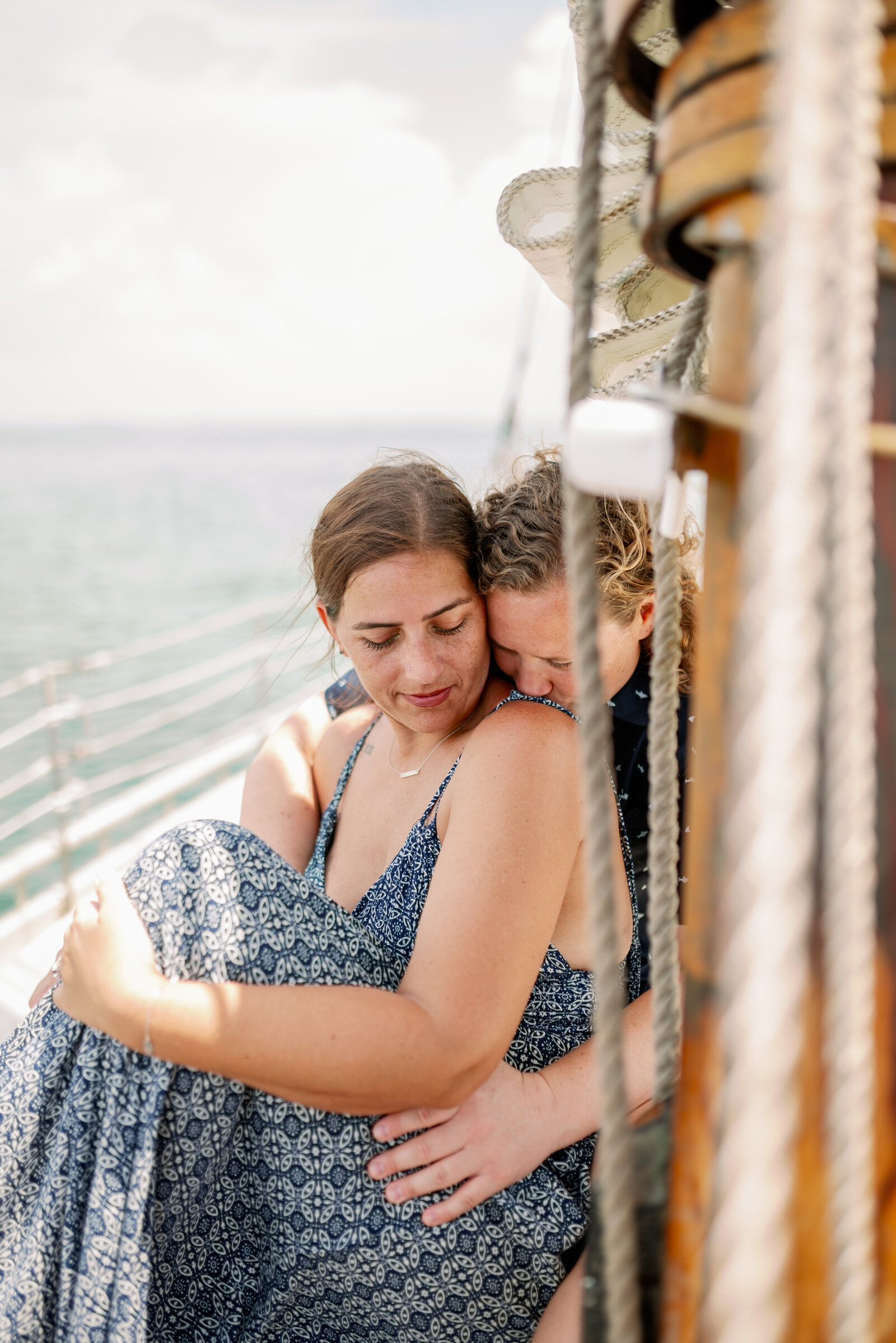 one women kissing the other on the shoulder, photographed from behind a sailboat rigging