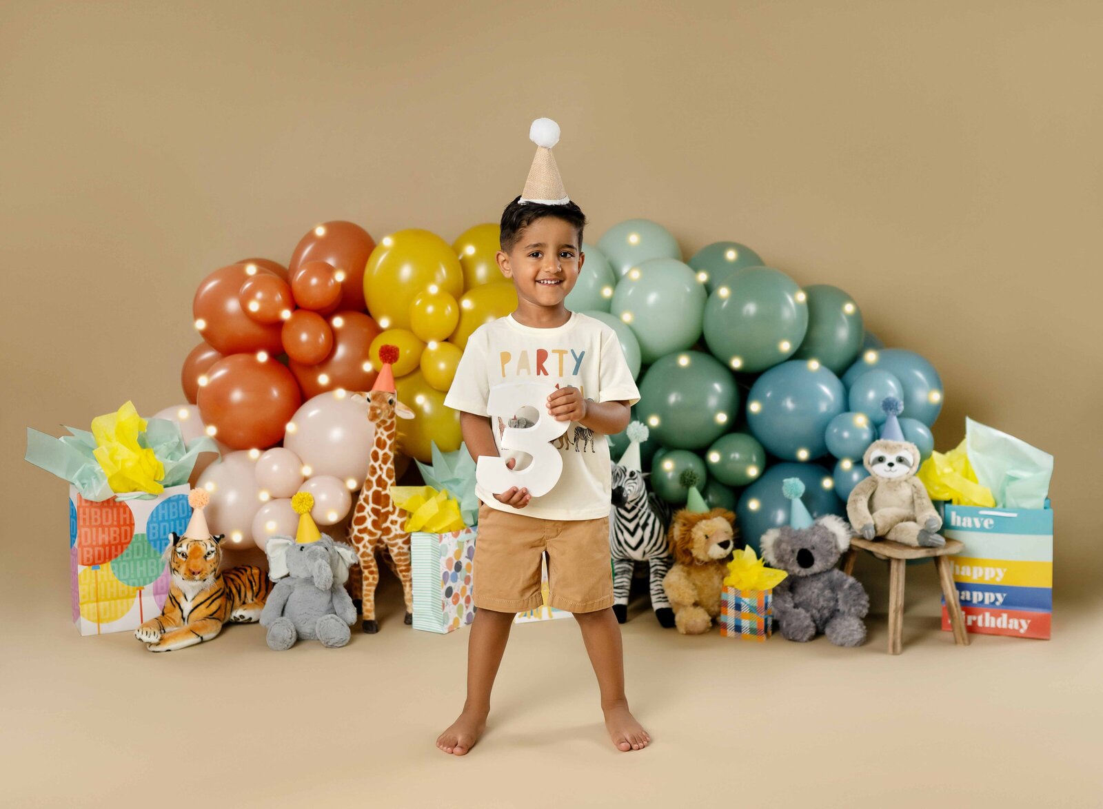 Party animal birthday set with retro balloon garland and 3 year old boy standing and holding #3, smiling at camera.