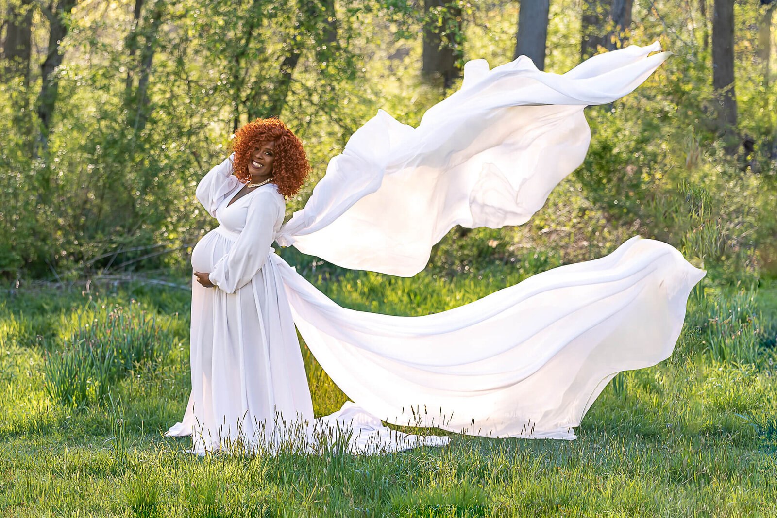 A woman in a flowing white gown stands in a green meadow, her dress billowing behind her like angelic wings, illuminated by the sunlight.